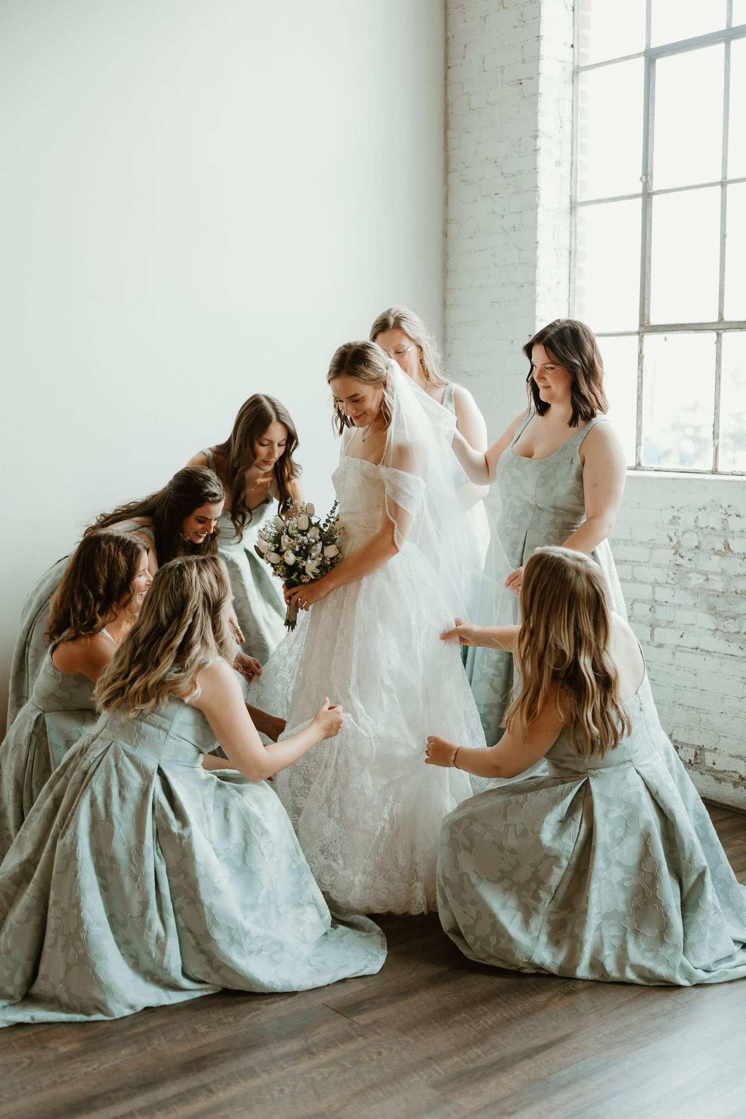 A bride in white and bridesmaids in sage green dresses pose playfully in a bright studio with white brick walls.