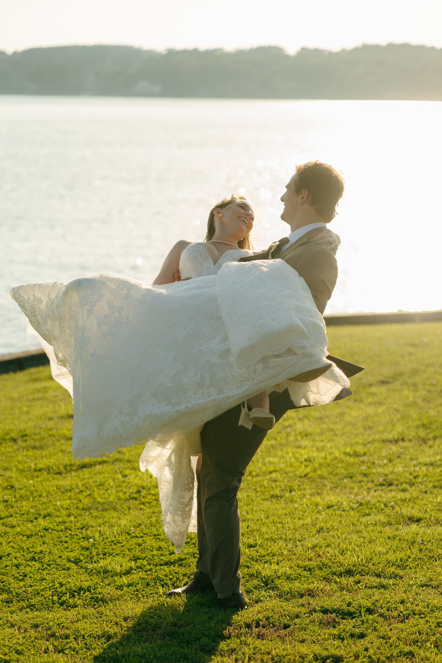 Groom lifting bride in white gown by waterfront at golden hour on green lawn.