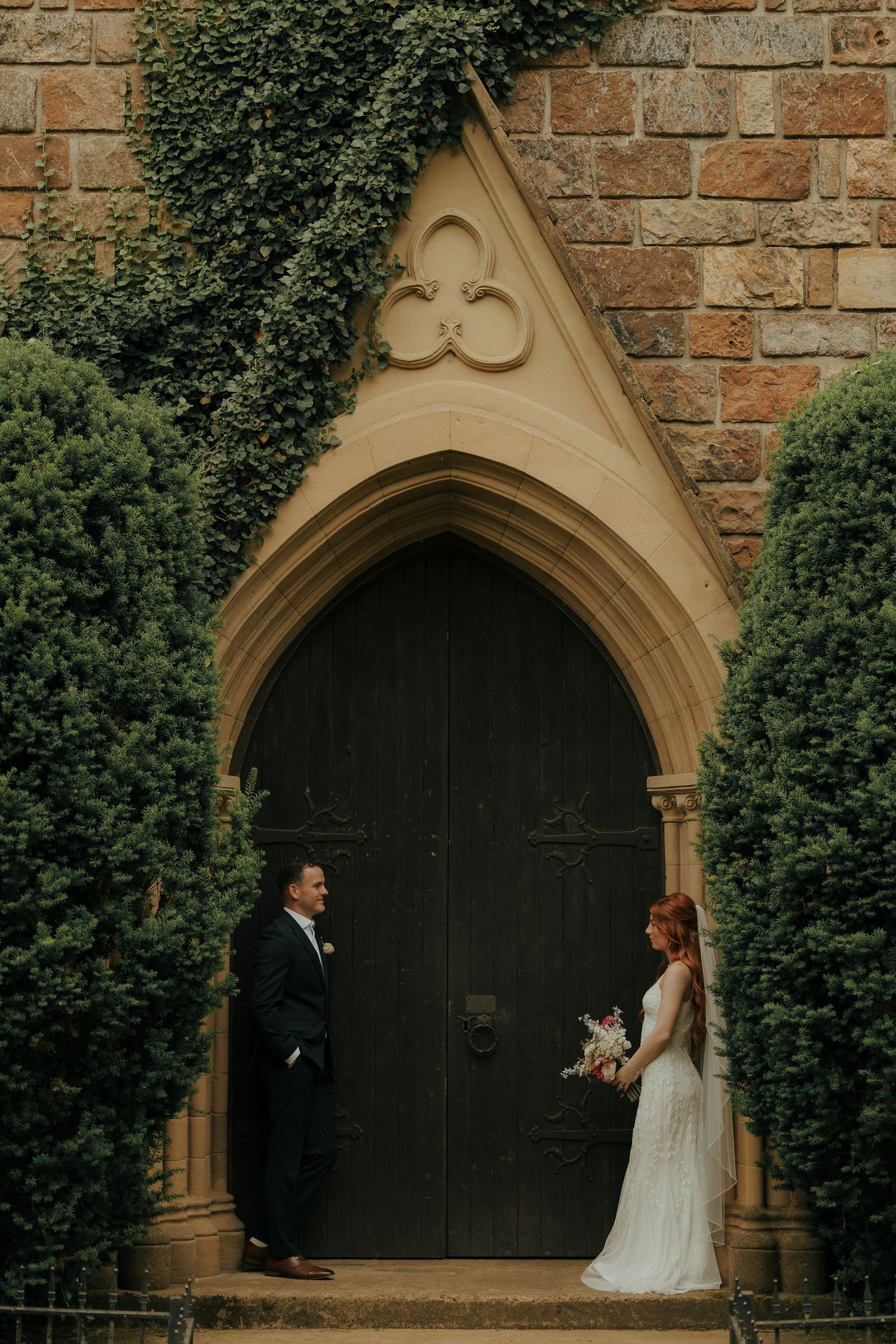 Bride and groom stand facing each other before a Gothic arched wooden door surrounded by lush green hedges at a stone church.