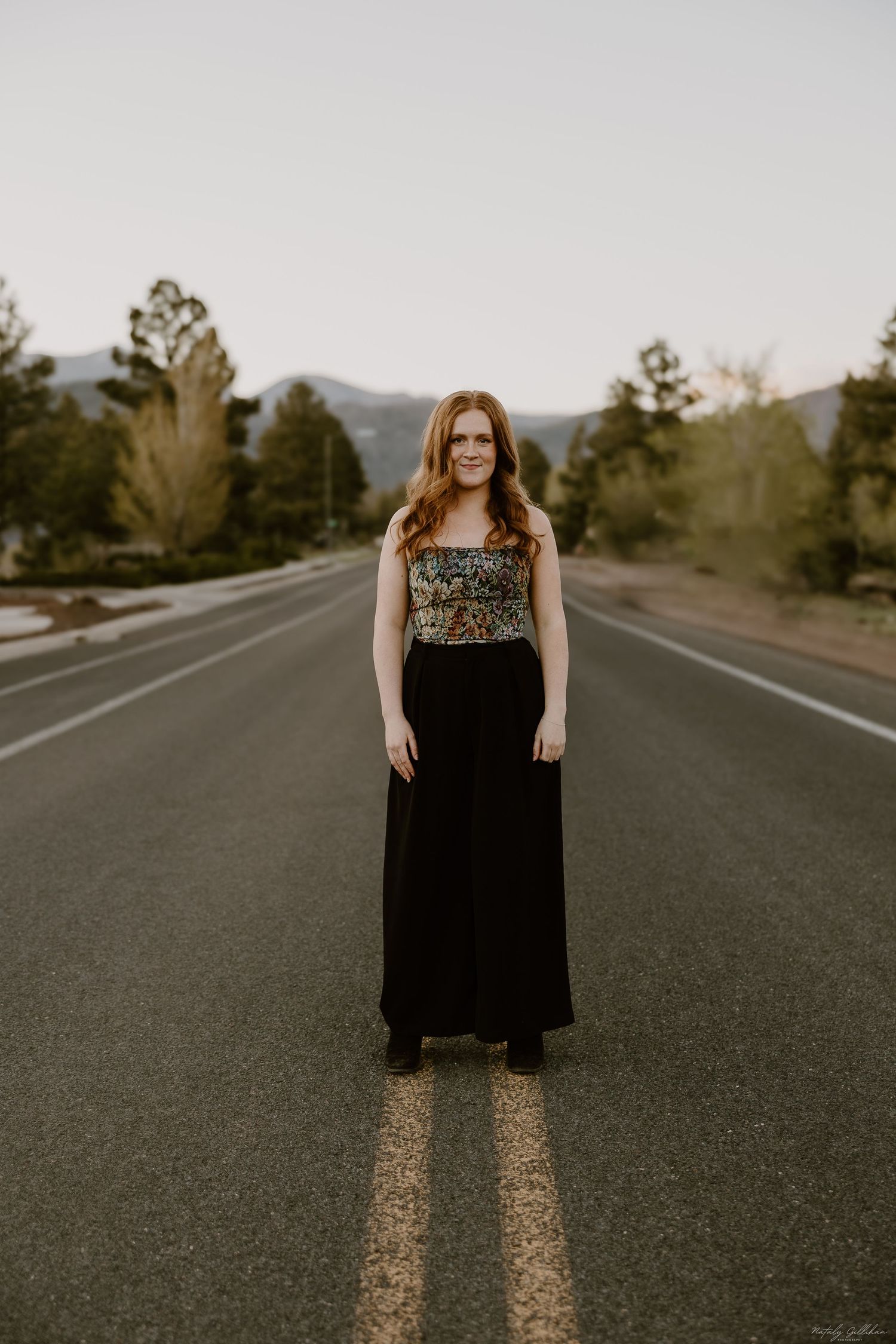 A woman stands in the middle of an empty mountain road at sunset wearing a black maxi dress with embellished bodice.