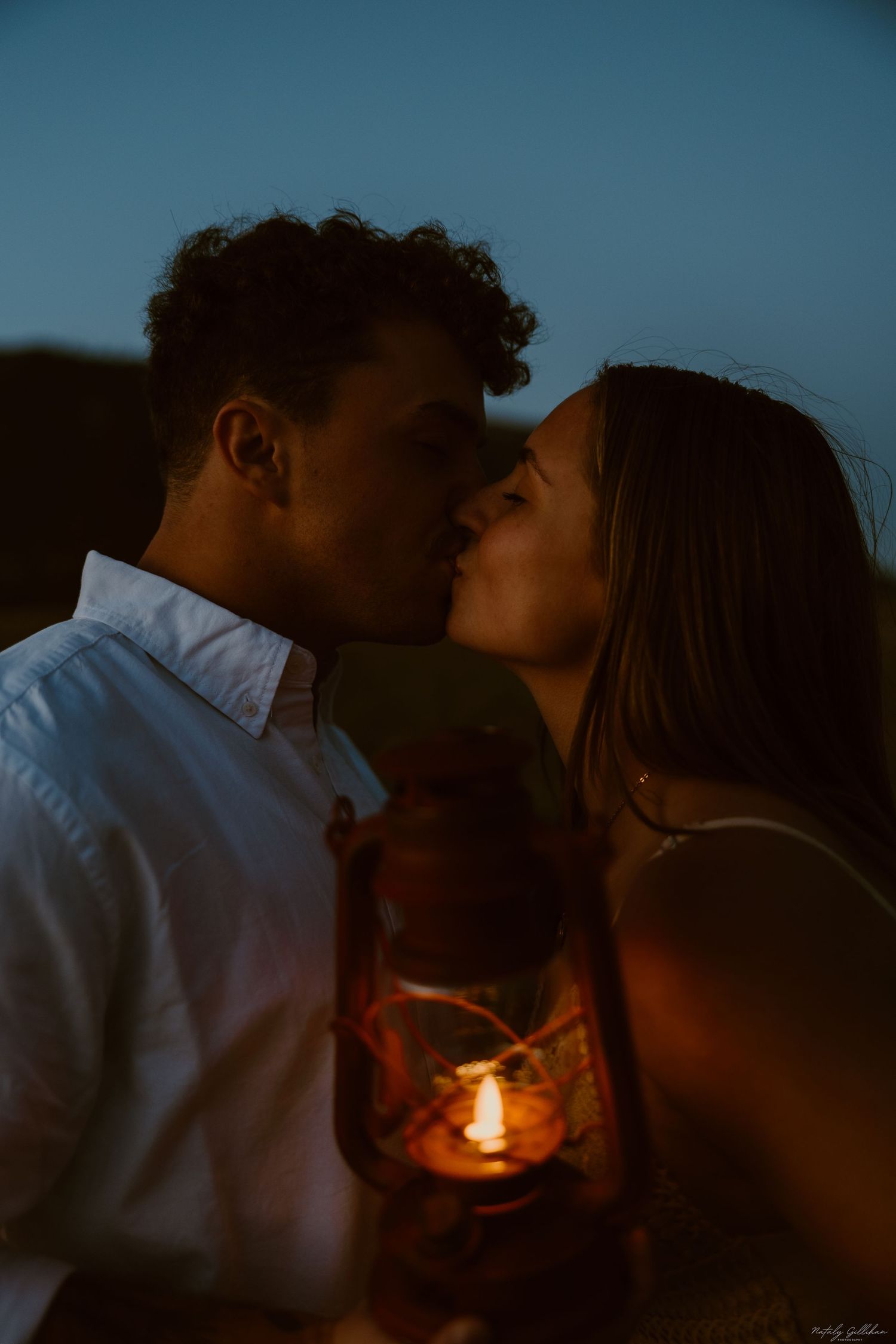 A romantic candlelit scene of a couple sharing an intimate kiss at dusk with a lantern glowing between them.