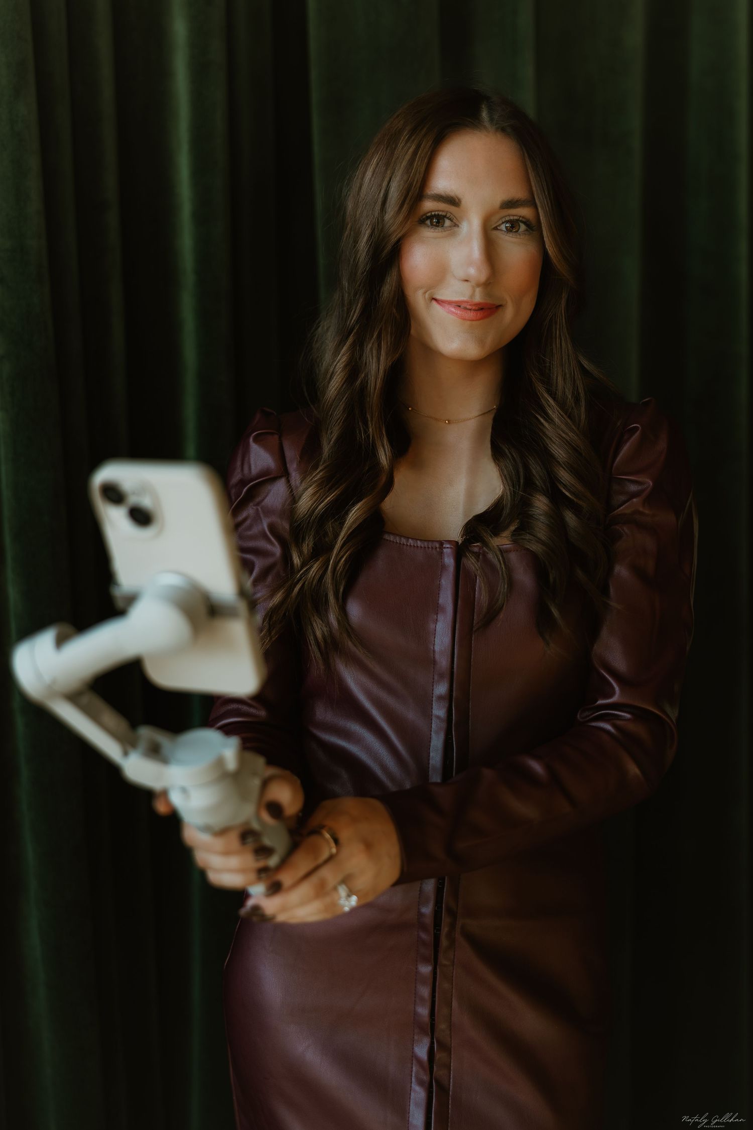 A person wearing a brown leather dress holds a white iPhone against a dark green curtain backdrop.