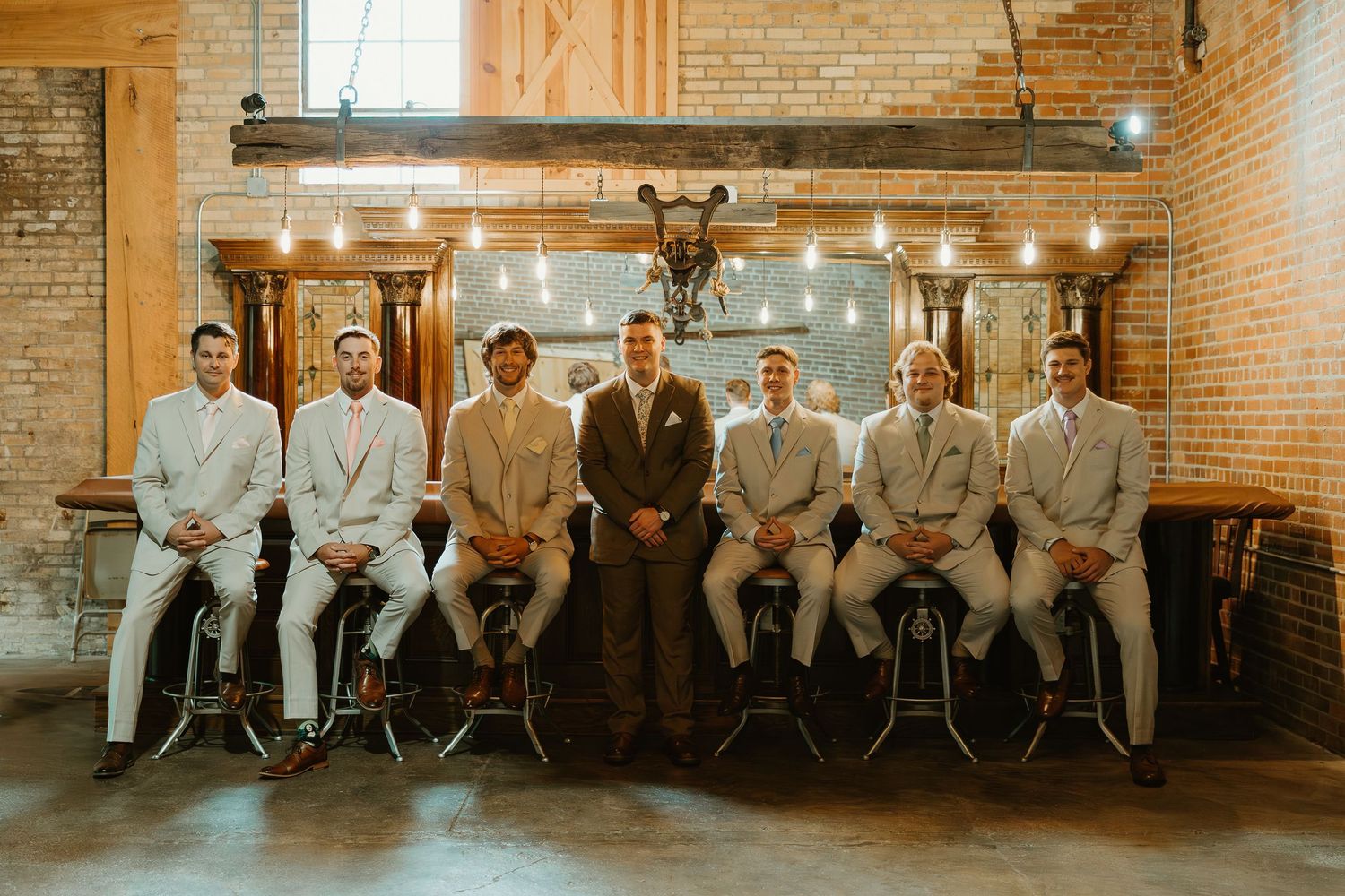 A group in neutral toned attire sits together inside a rustic brick-walled venue with warm lighting.