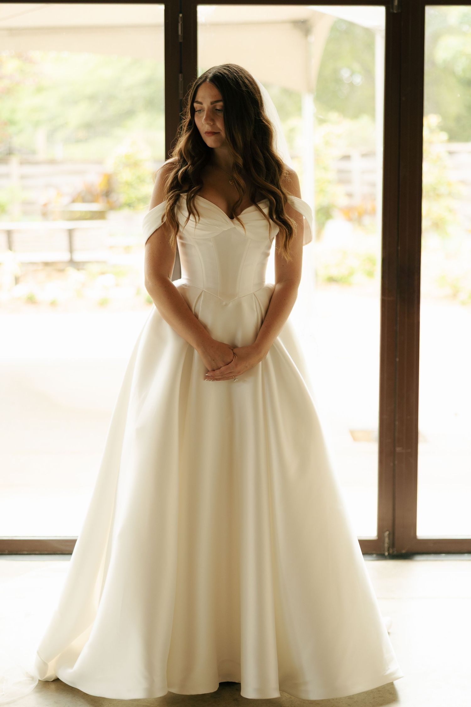 Bride wearing a classic ivory off-shoulder A-line wedding gown with sweetheart neckline, standing in soft natural light.
