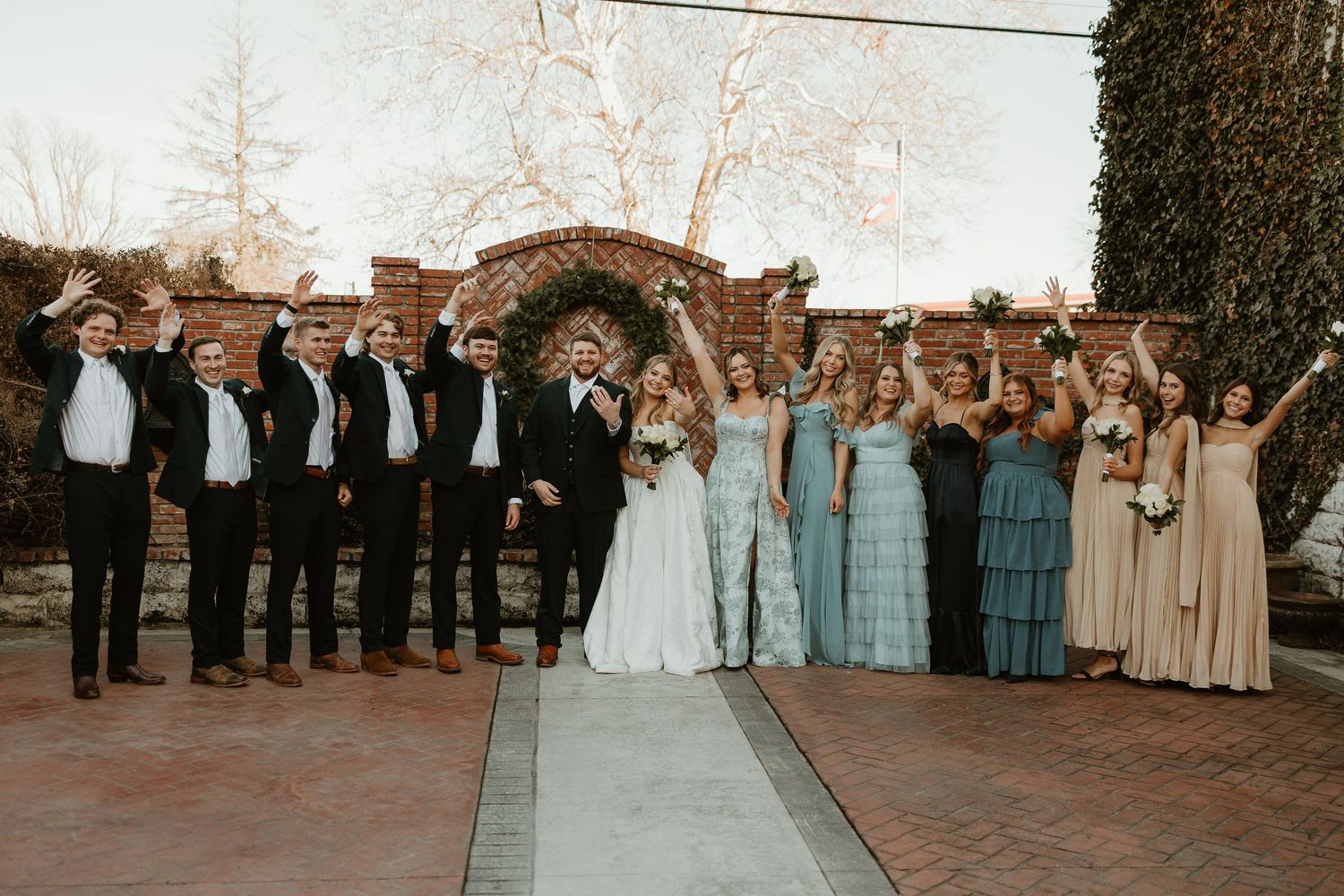 Wedding party in formal attire poses together under rustic arch with bridesmaids in blue and beige dresses.