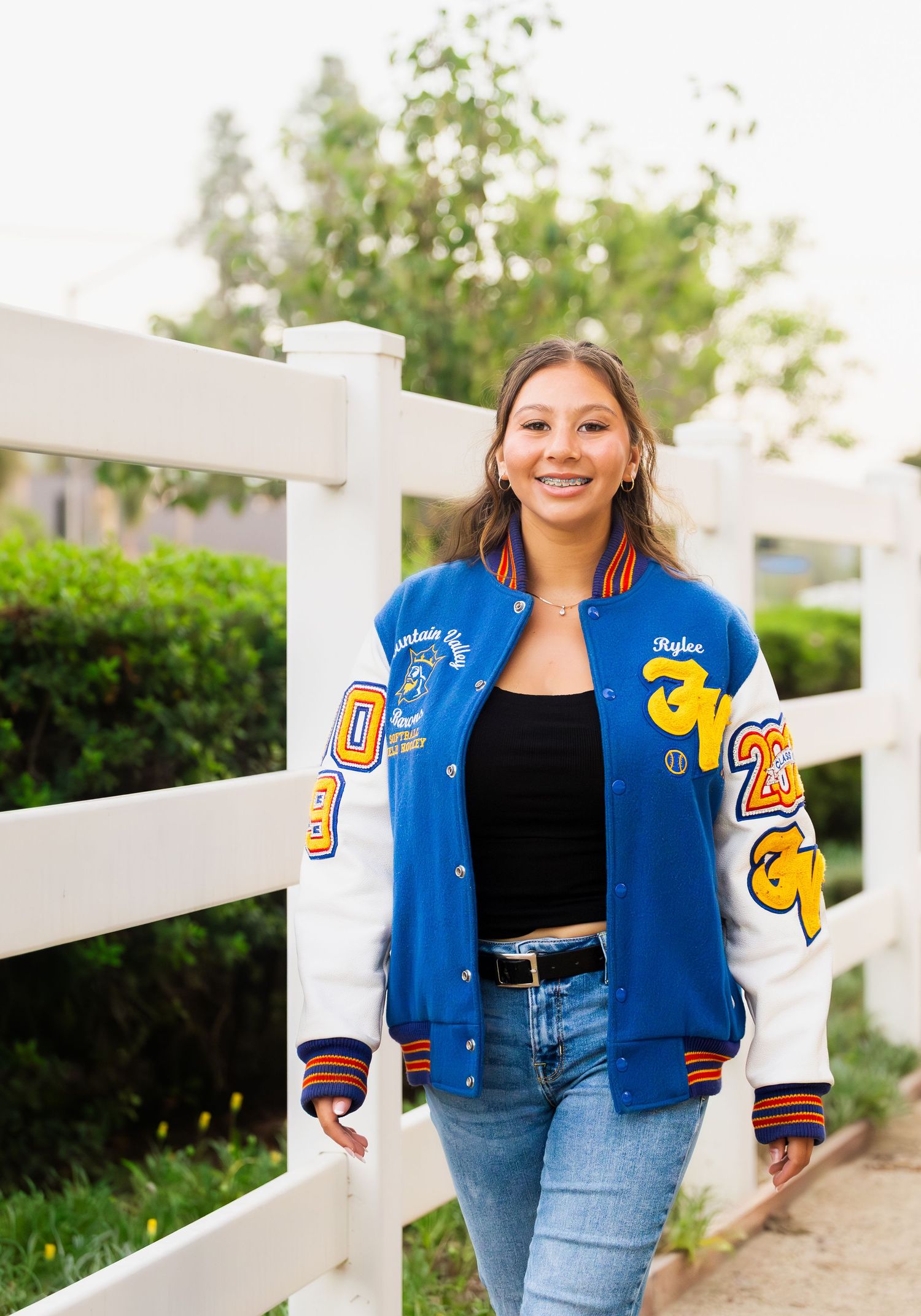 A varsity letterman jacket in royal blue and gold paired with denim jeans and a black top against a white fence backdrop.
