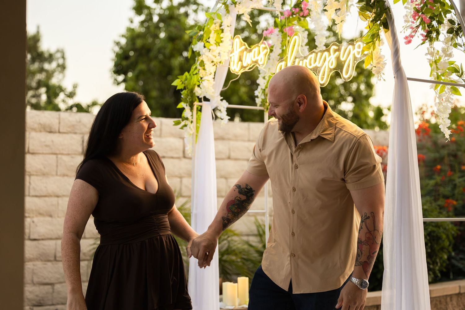 Couple holding hands and smiling under a floral wedding arch with neon sign at an outdoor ceremony.