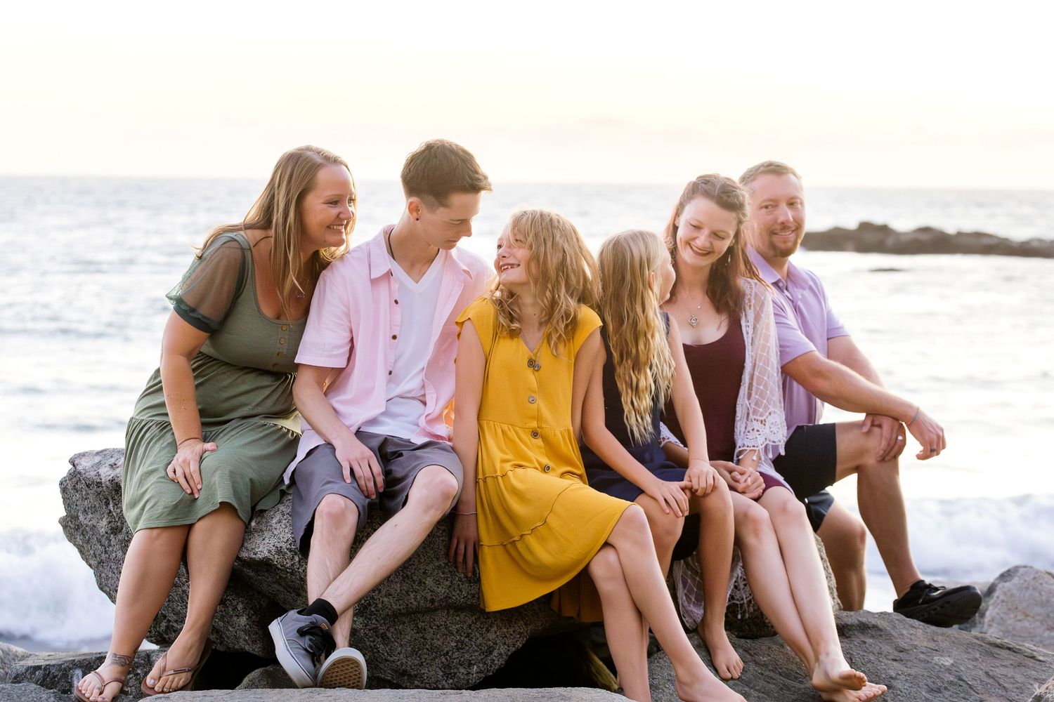 Group of friends sitting together and laughing on a rocky beach at sunset with ocean waves in background.