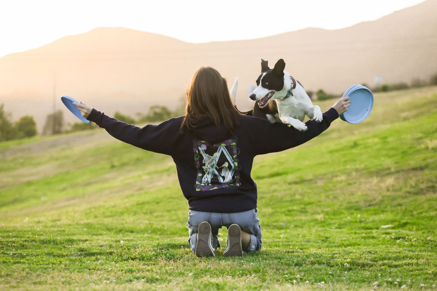 Woman kneeling on grass with arms outstretched, balancing a dog on her back against a scenic mountain sunset.