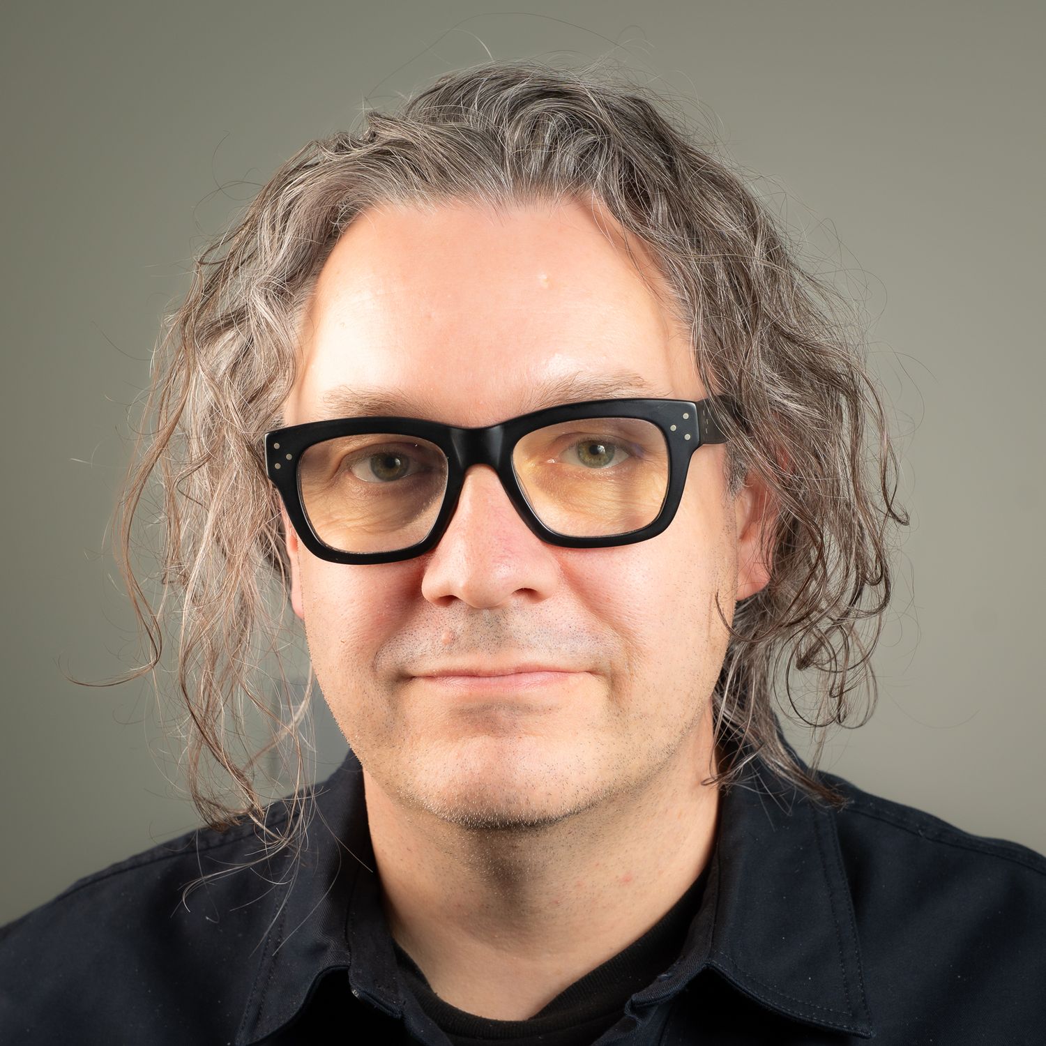 Keith with long brown hair and black thick-rimmed glasses wearing a dark shirt, professional headshot on gray background.