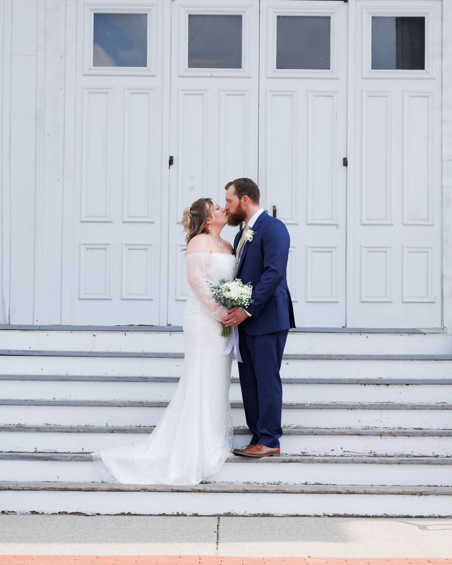 A couple sharing a romantic moment on white church steps at their outdoor wedding ceremony.