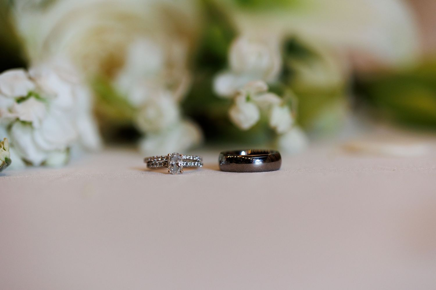 Wedding rings rest on a light pink surface with delicate white flowers creating a romantic backdrop.