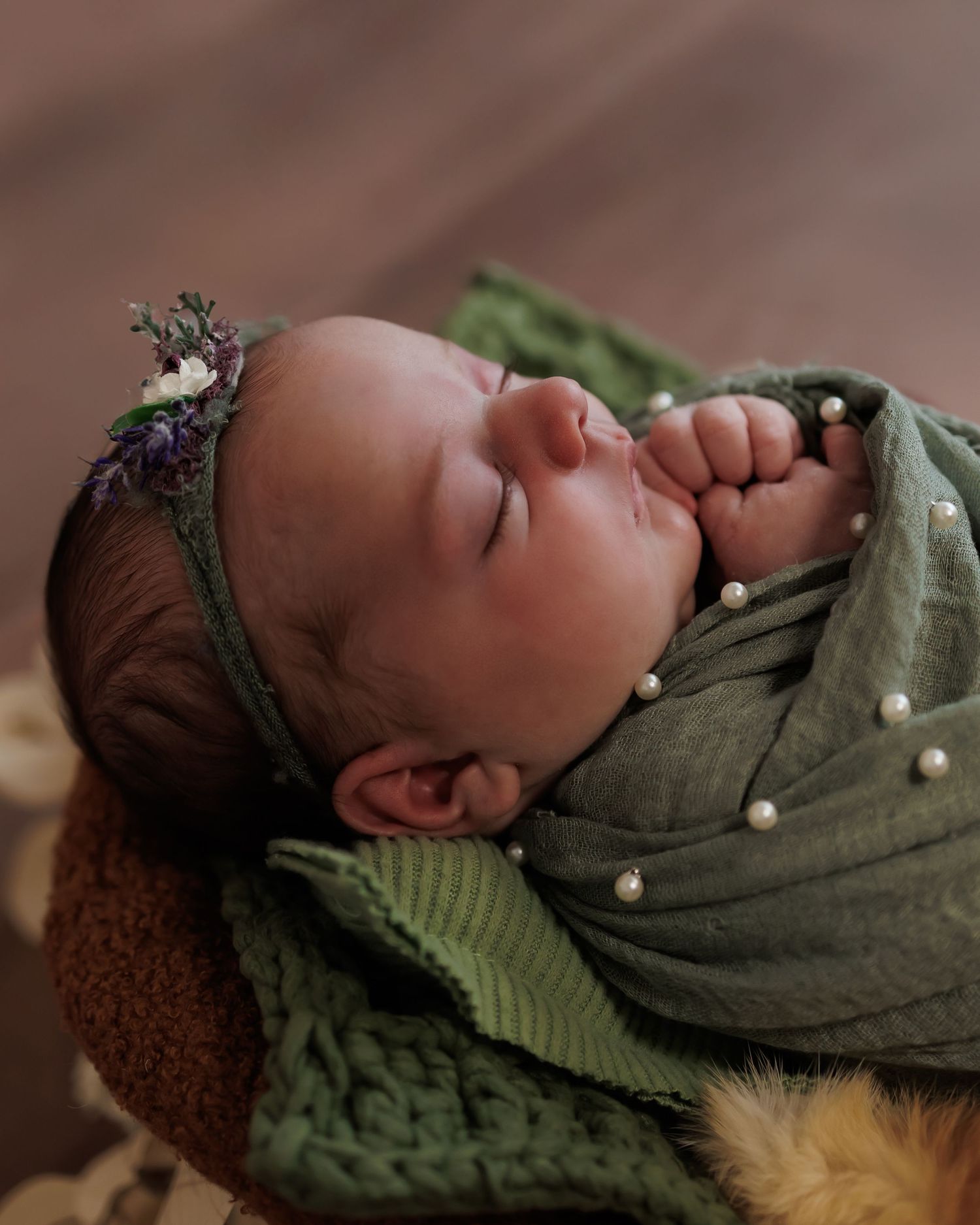 A newborn baby sleeps peacefully wrapped in an olive green swaddle blanket against a brown textured backdrop.
