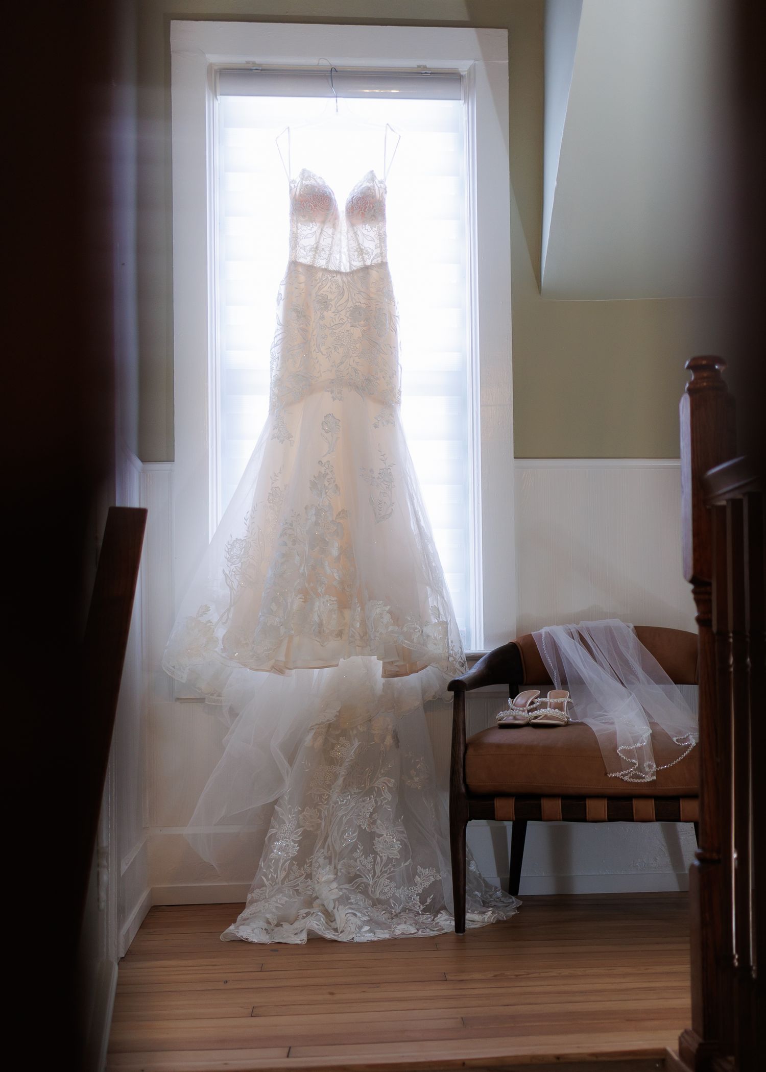 A white lace wedding dress hangs in front of a sunlit window creating an ethereal silhouette.