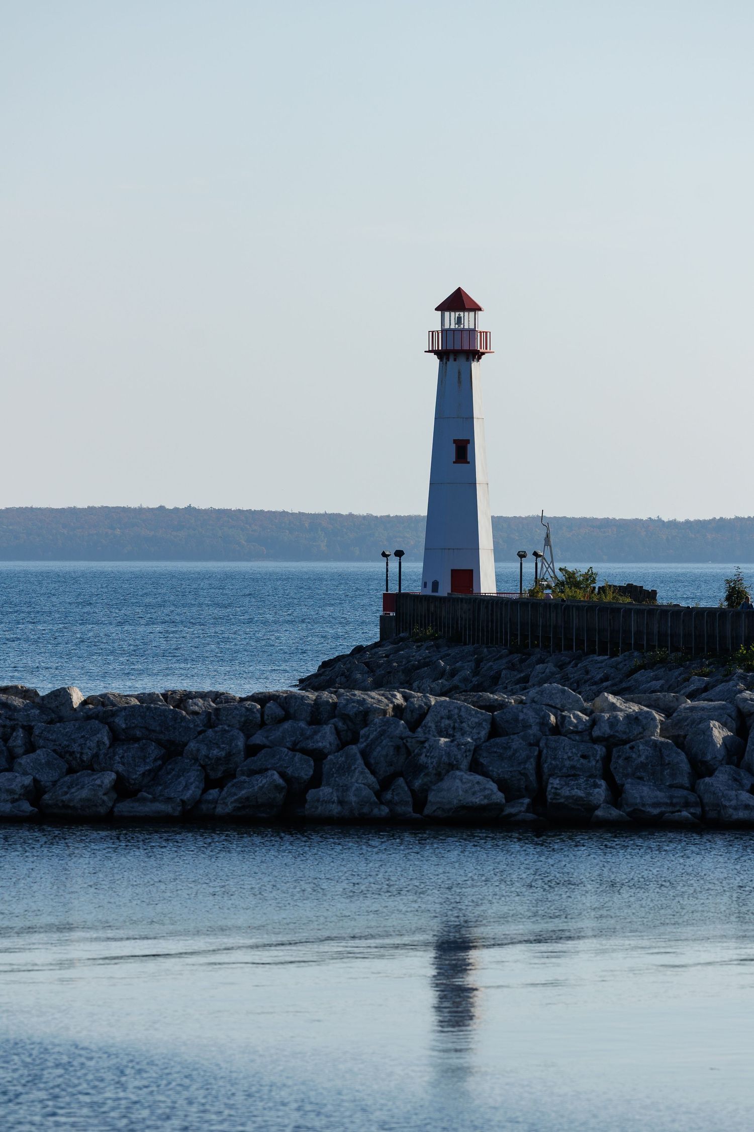 Lighthouse on rocky breakwater with calm blue waters and distant shoreline at dusk.