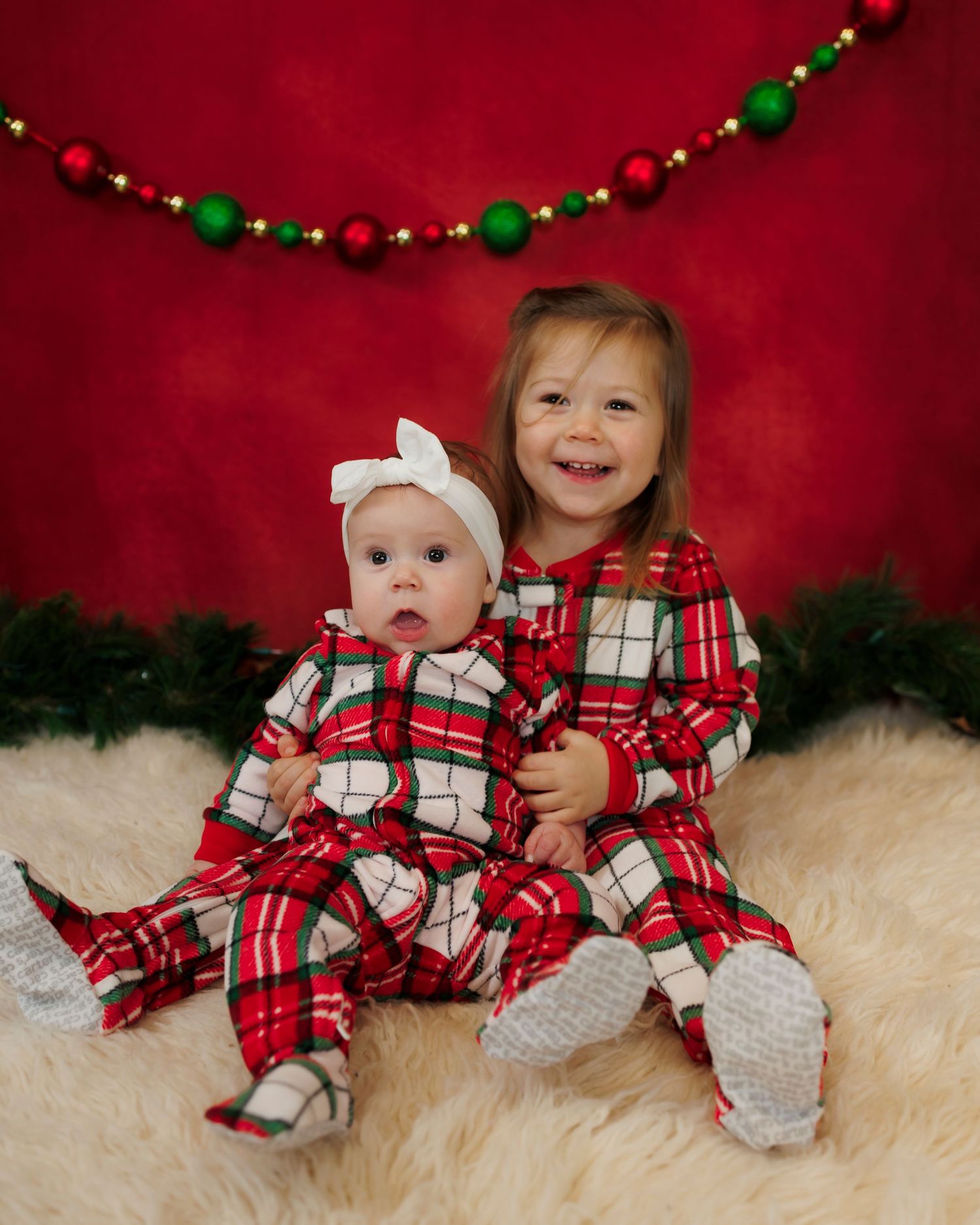 Two children wearing matching red plaid Christmas pajamas sitting on white fur against a red background with holiday decor.