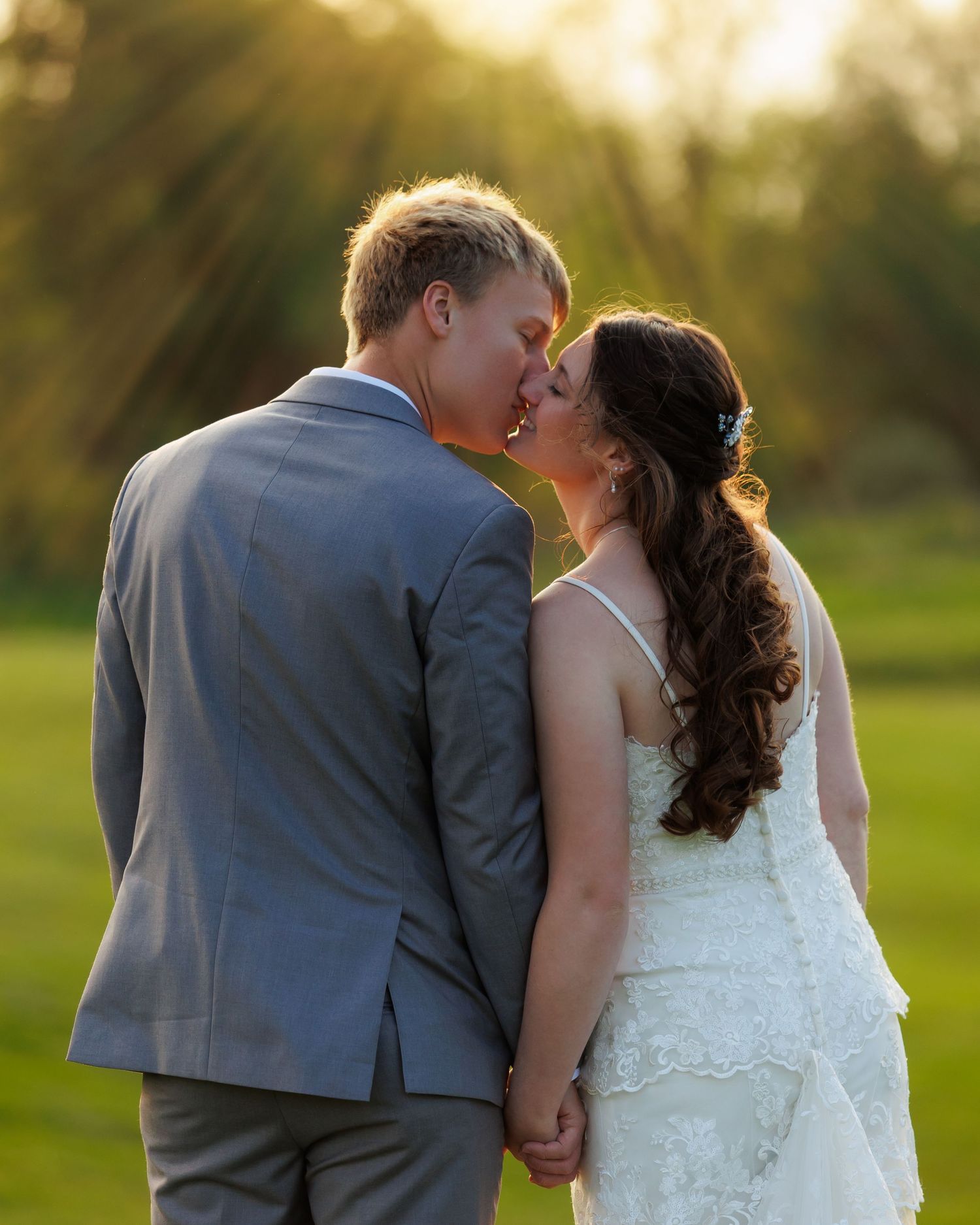 A couple in wedding attire share romantic kisses during their outdoor sunset photo session in a grassy field.