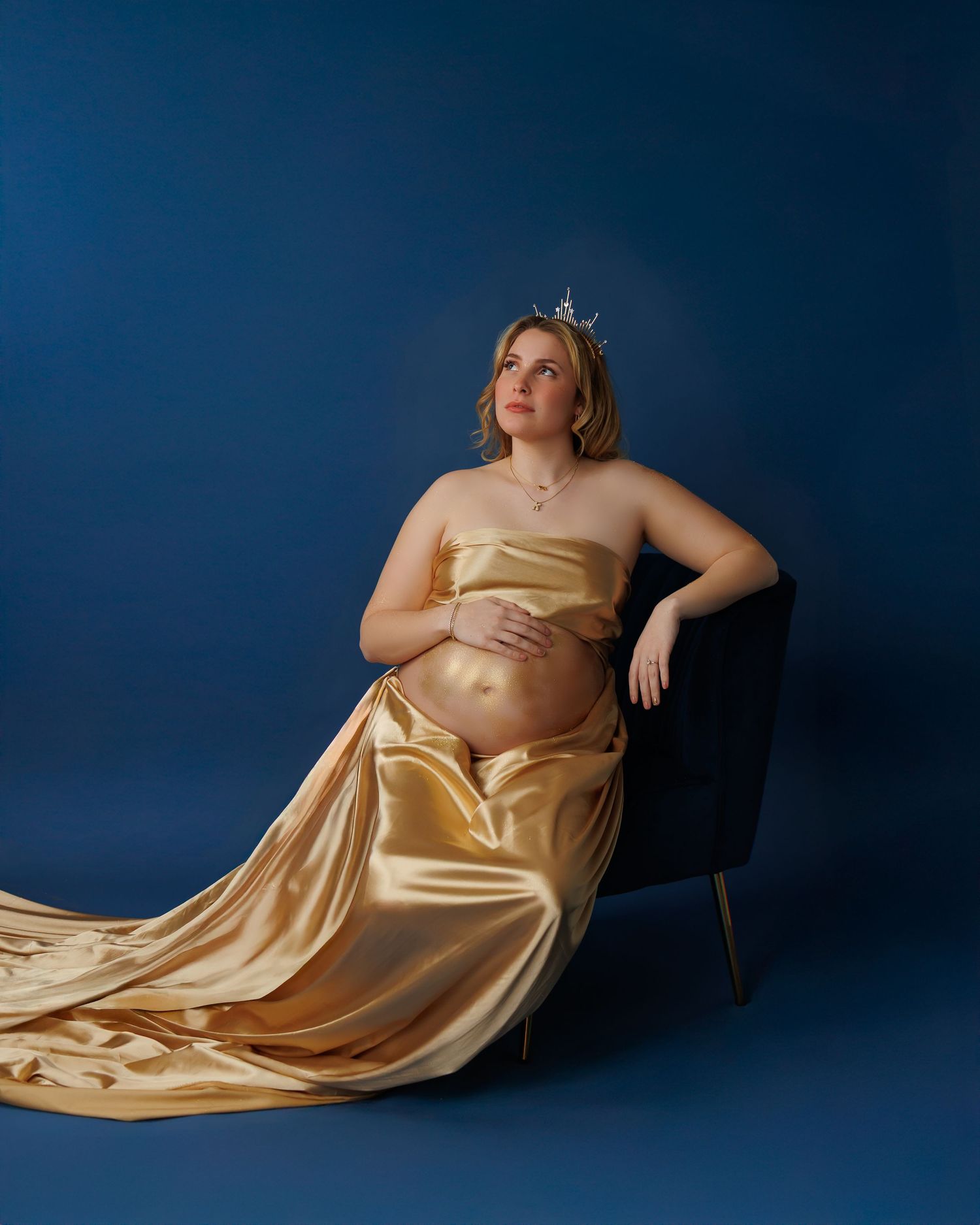Artistic maternity photoshoot featuring flowing gold silk dress against dark blue backdrop in studio setting.