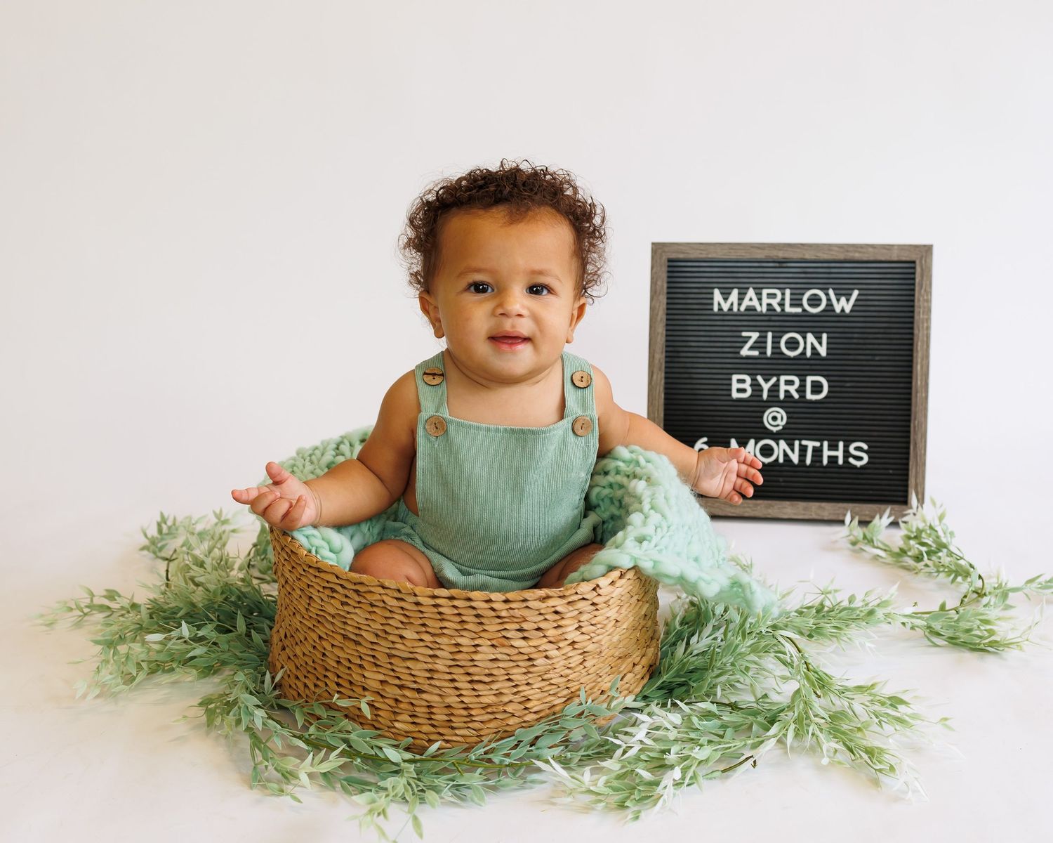 Baby in green romper sits in woven basket with greenery and letterboard displaying age milestone.