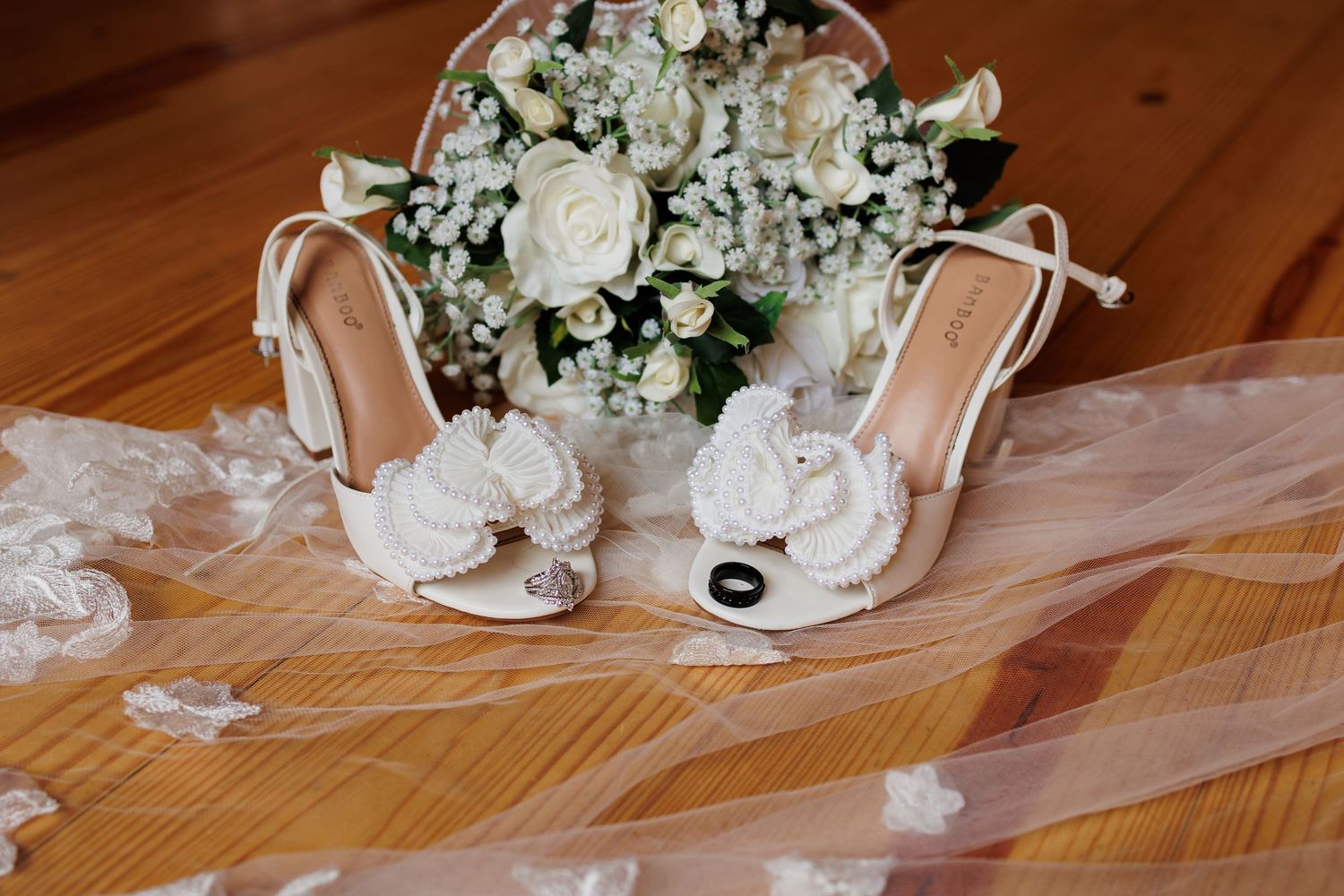 White wedding shoes and delicate floral bouquet arranged on wooden floor with flowing veil.