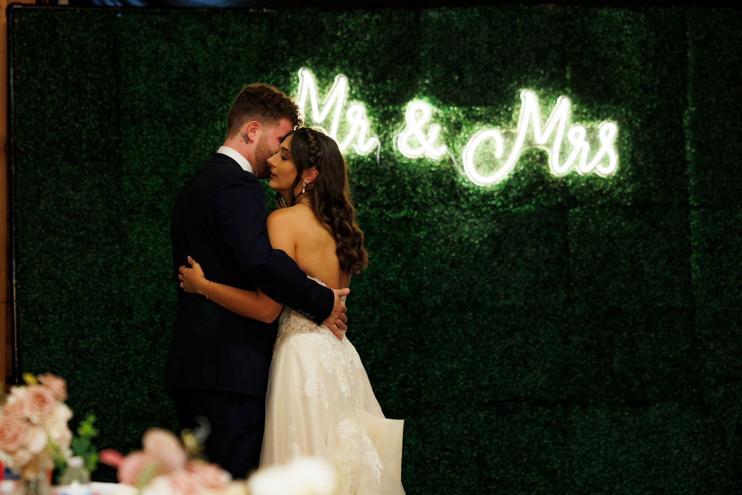 A couple shares a romantic kiss on their wedding day with a neon sign glowing in the background.