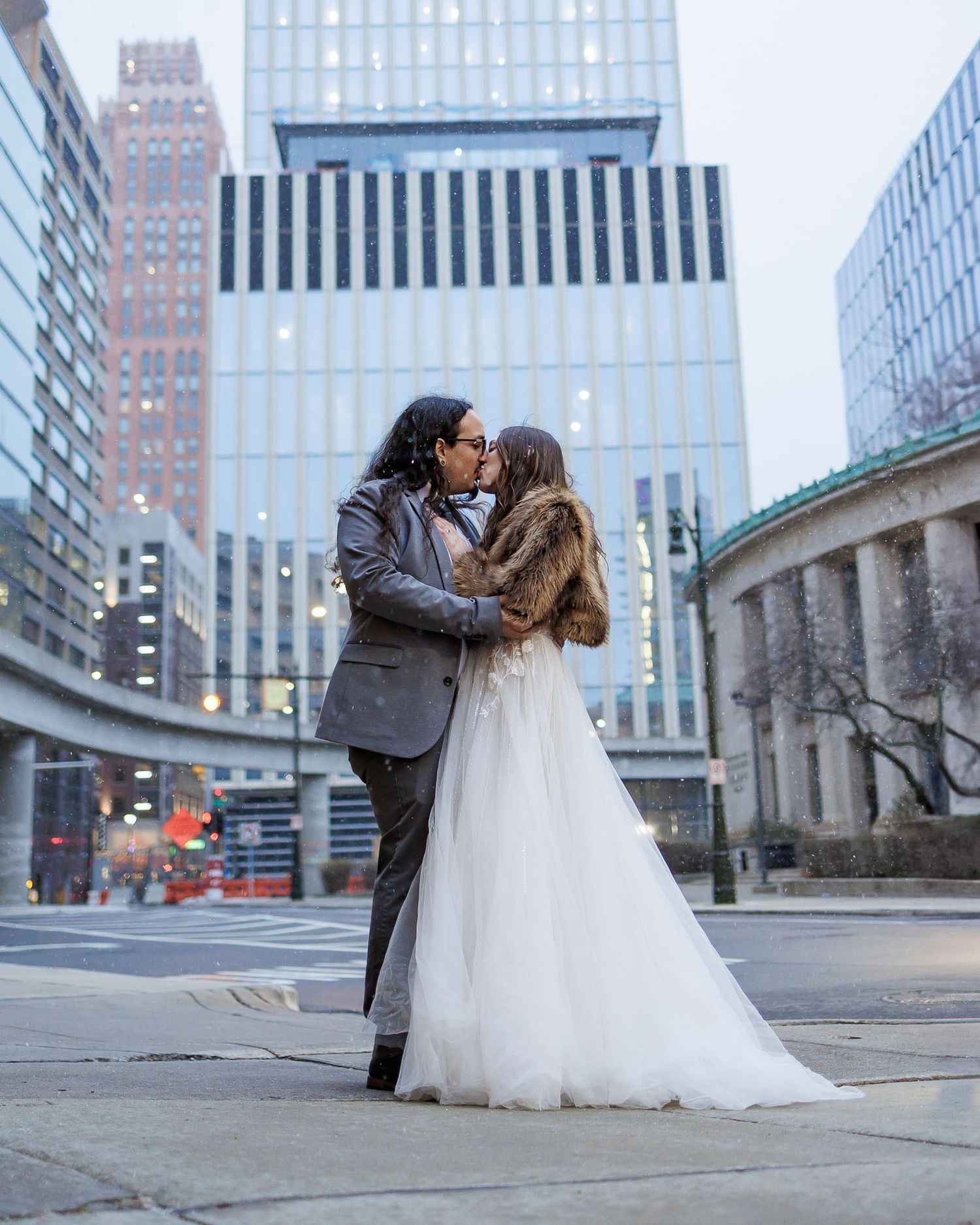 A romantic couple in wedding attire share intimate moments against an urban cityscape backdrop with tall buildings.