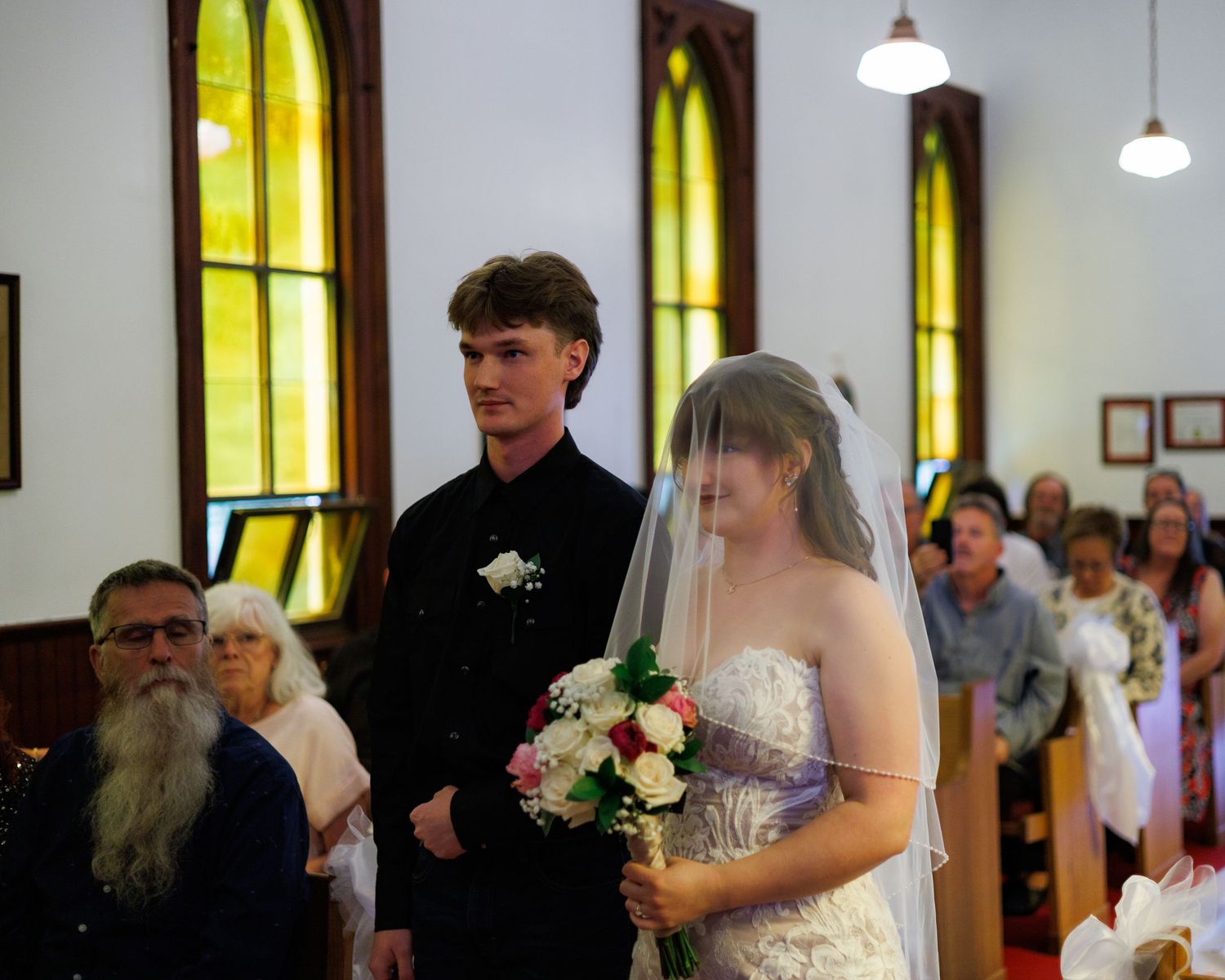 A couple stands at the altar during their wedding ceremony in a church with yellow stained glass windows.