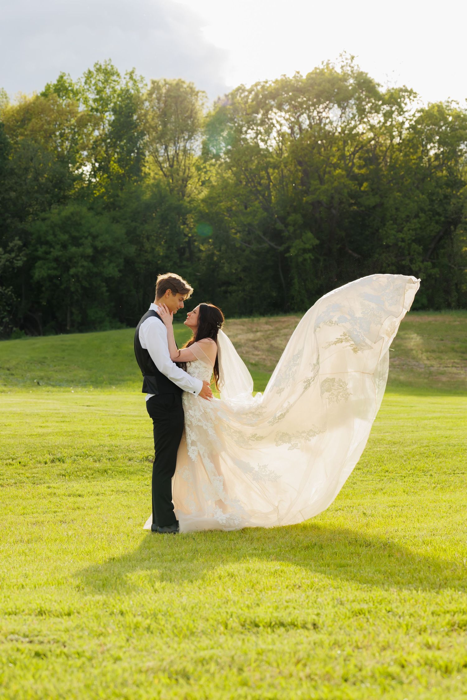 A romantic wedding photo in a sunlit field with the bride's flowing white dress veil caught dramatically in the breeze.