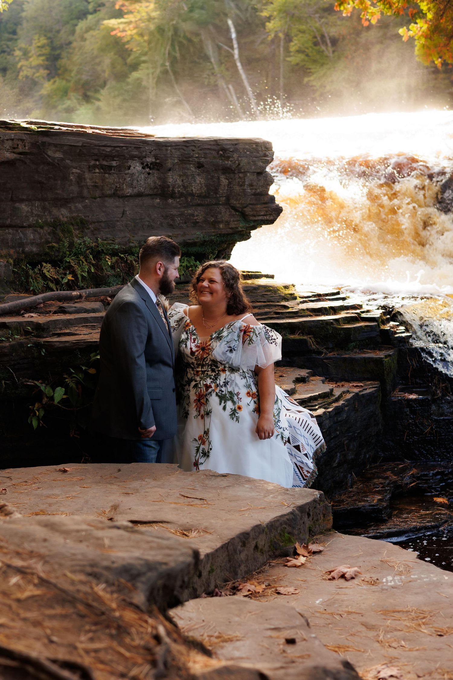 A couple in wedding attire embraces near a rushing waterfall on rocky terrain during autumn.