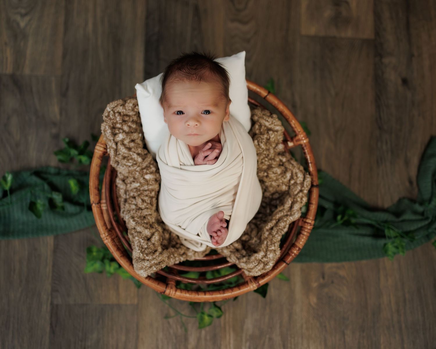 A baby dressed in white lies in a round wicker basket with brown blanket on wooden floor.
