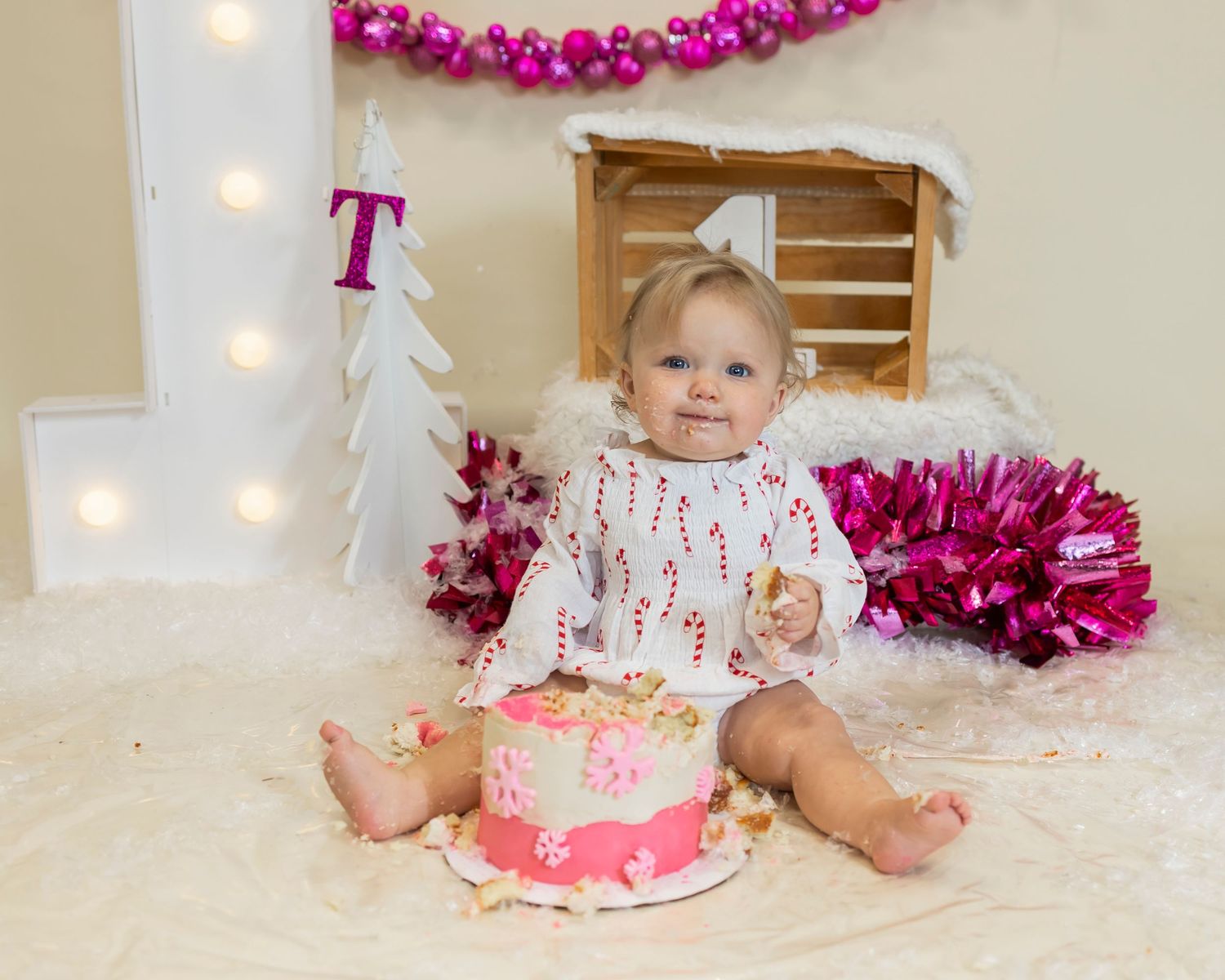 A baby in white clothing sits on a cream blanket with a pink birthday cake and magenta decorations in the background.
