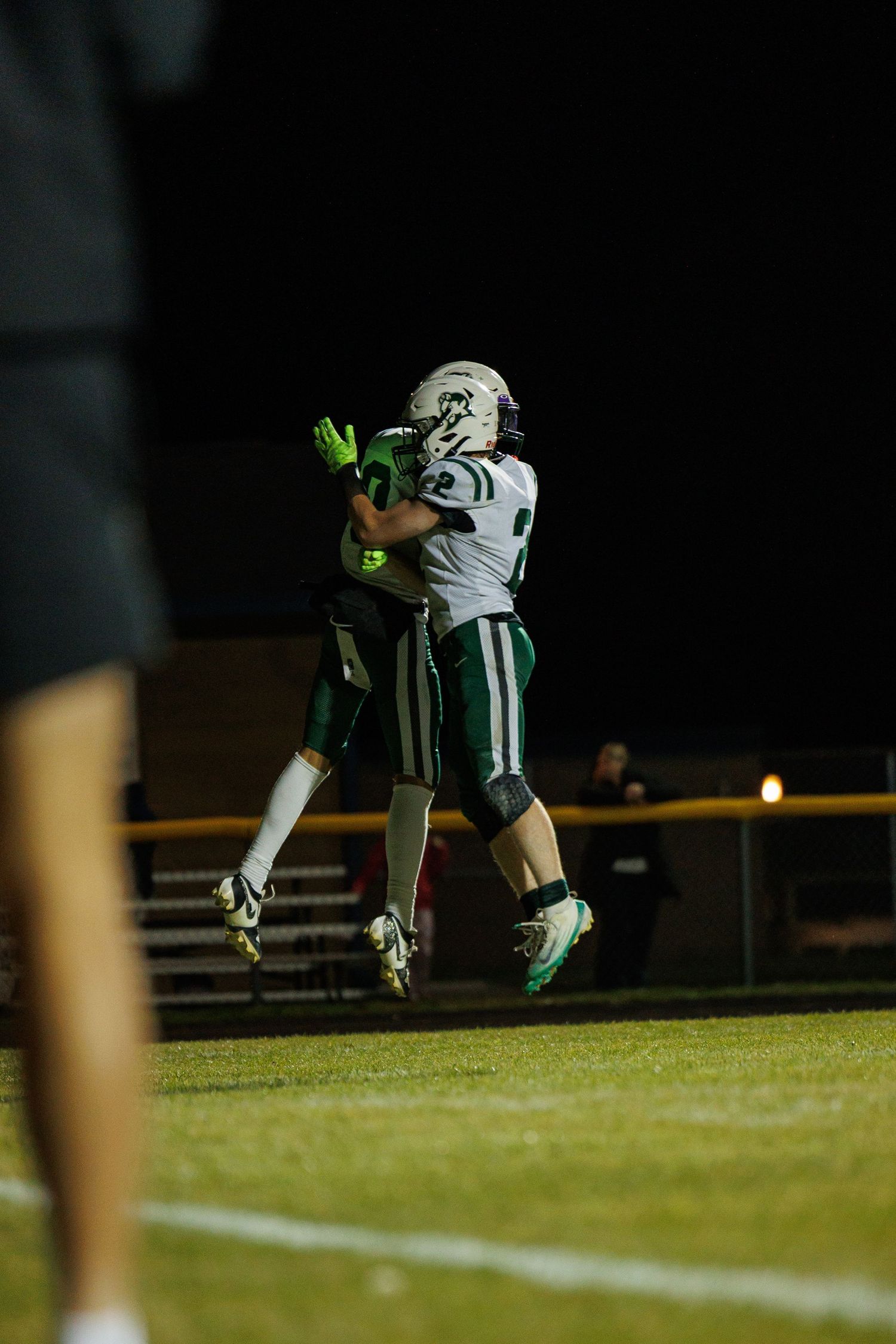 Football players in dark uniforms leap high to catch a pass during a nighttime game on an illuminated field.