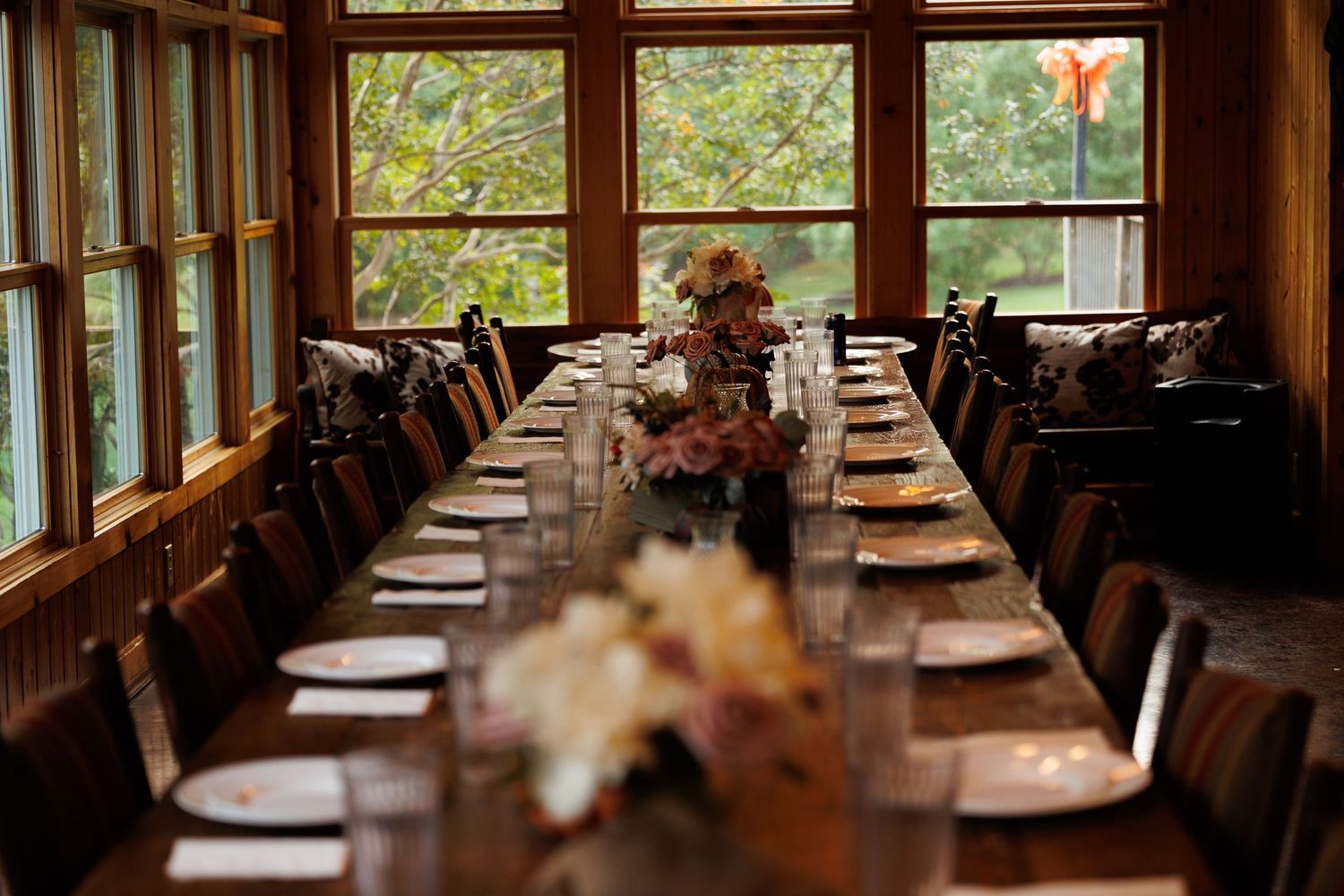Long wooden dining table set for an intimate gathering with fall floral centerpieces in a sunlit window-lined room.