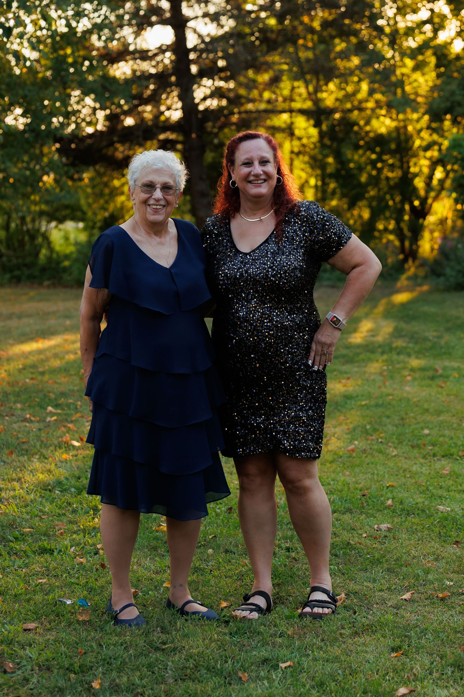 Two people pose together on a grassy lawn at sunset with golden light filtering through trees in the background.