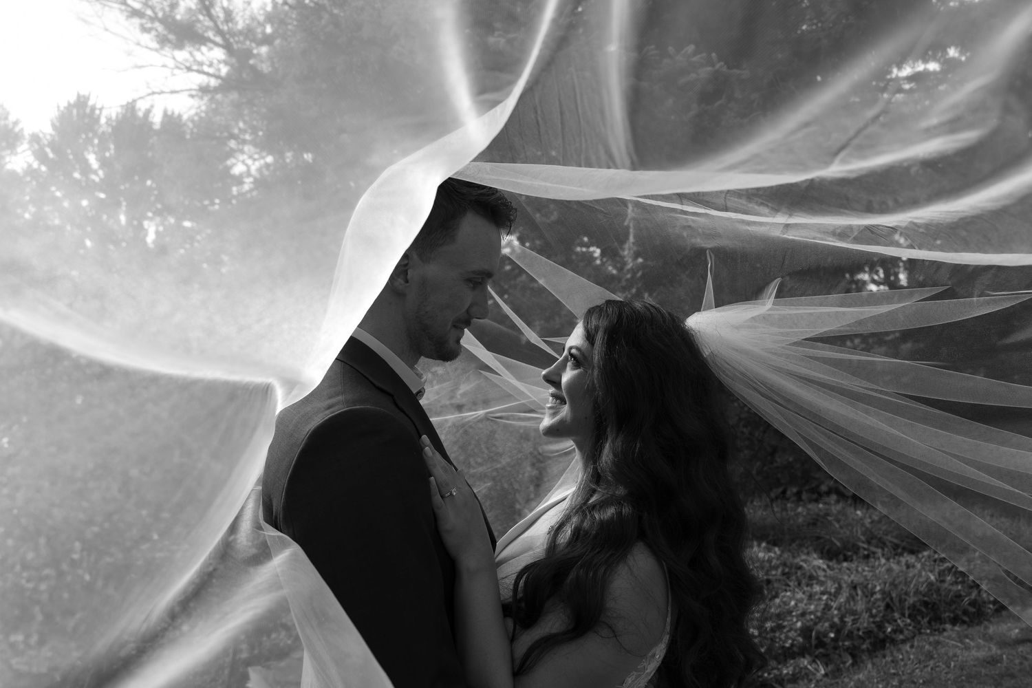 Ethereal black and white wedding photo showing a couple embracing under flowing veil fabric in an outdoor setting.