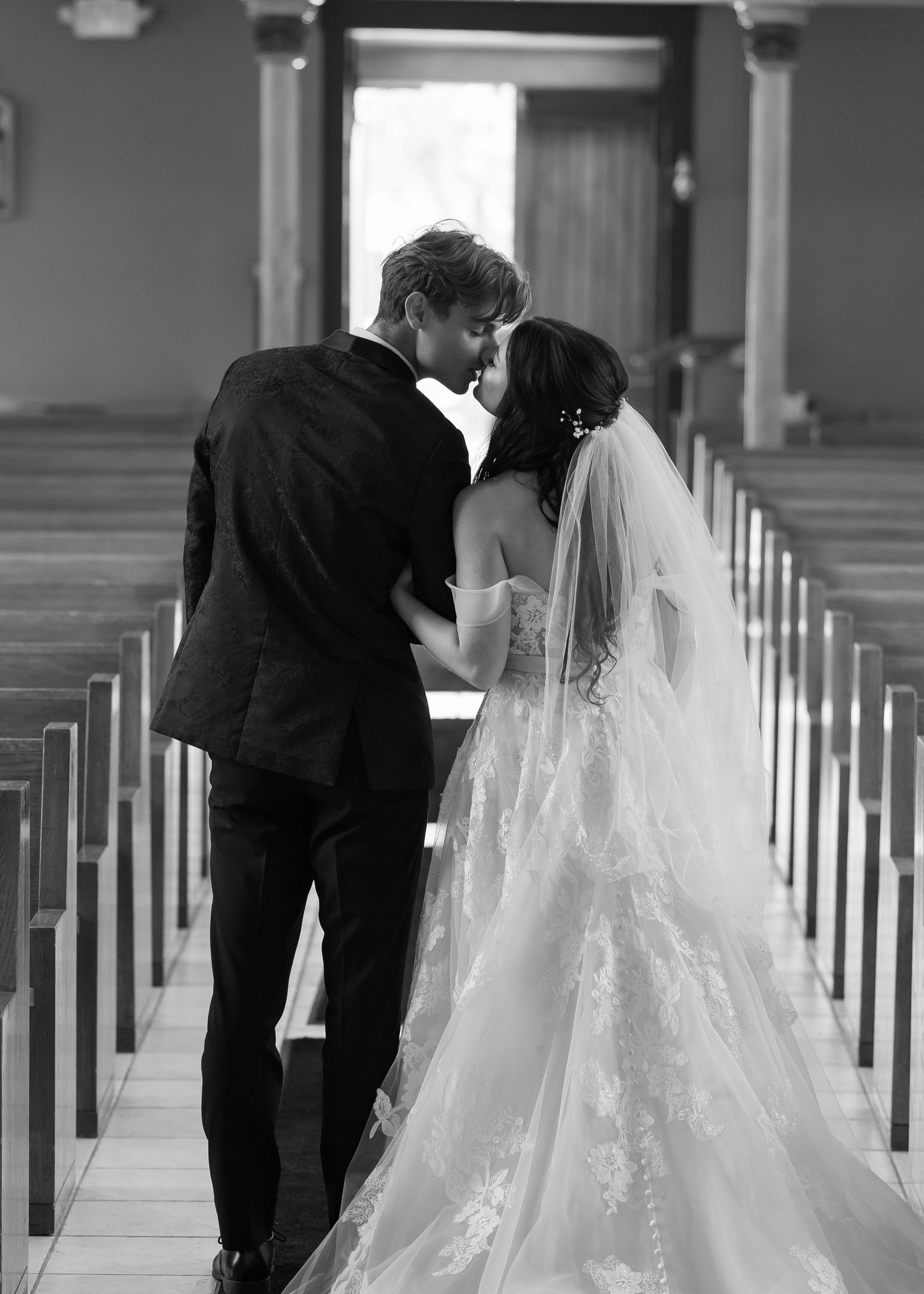 A couple shares their first kiss as newlyweds in a church aisle, with the bride wearing a flowing white veil and gown.