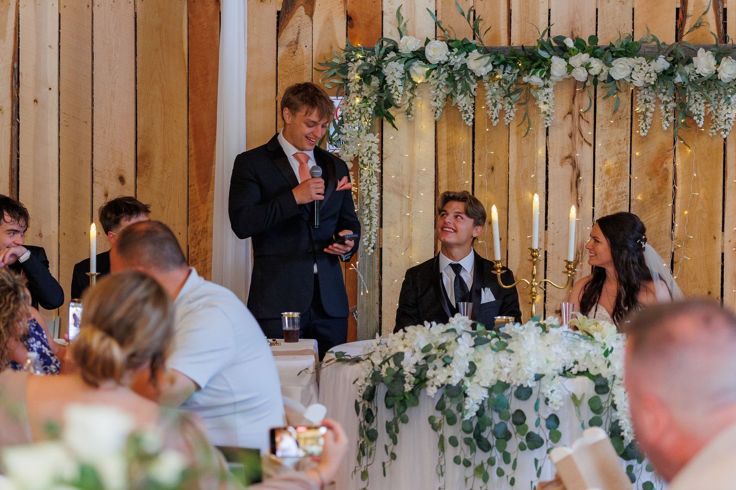 Wedding reception speech being delivered at rustic barn venue decorated with greenery garlands and white flowers.
