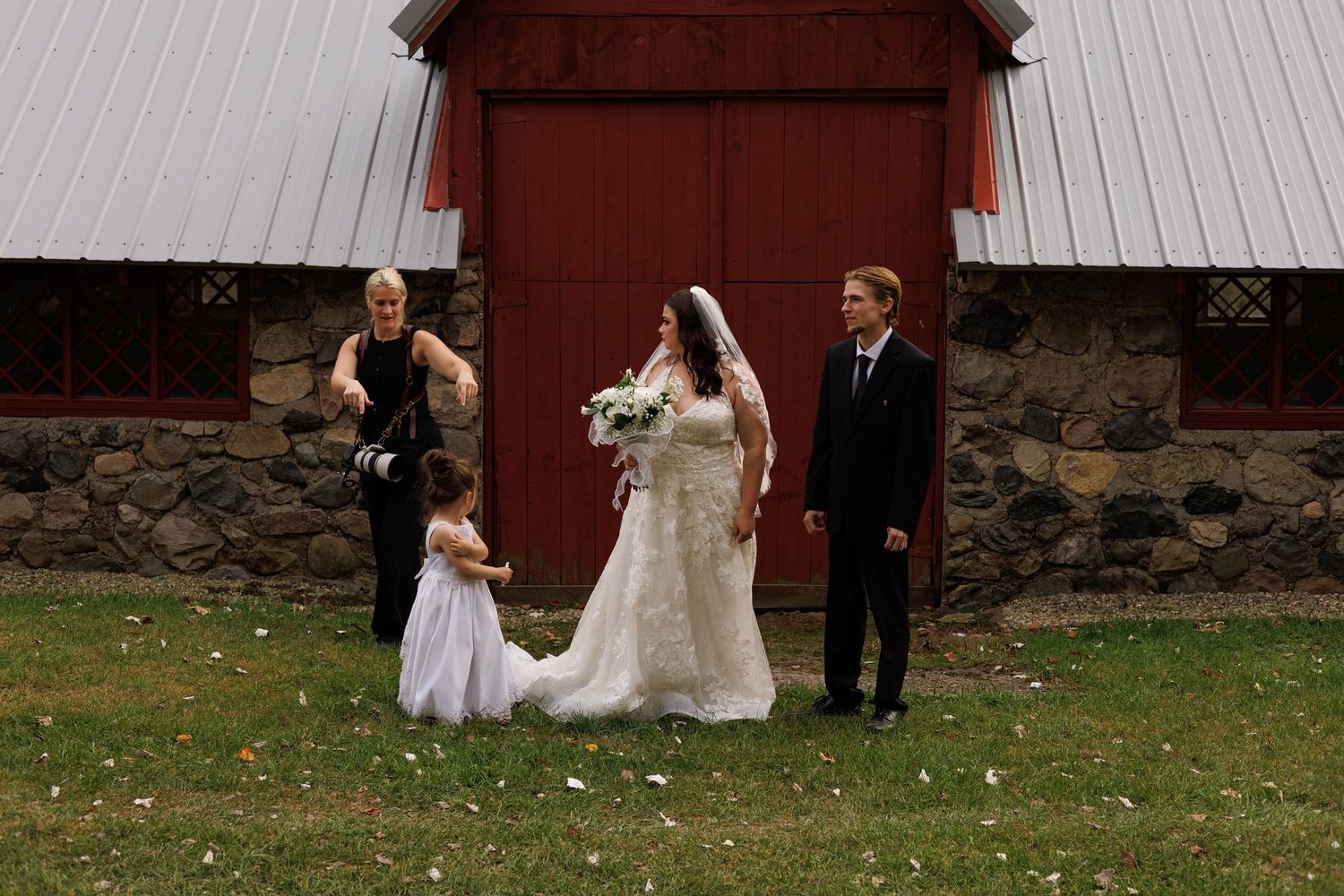 A wedding party walks in front of a rustic red barn and stone wall on an autumn day.