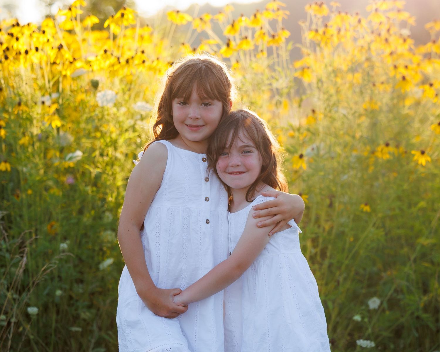 Two children in white dresses embrace while standing in a field of golden wildflowers at sunset.