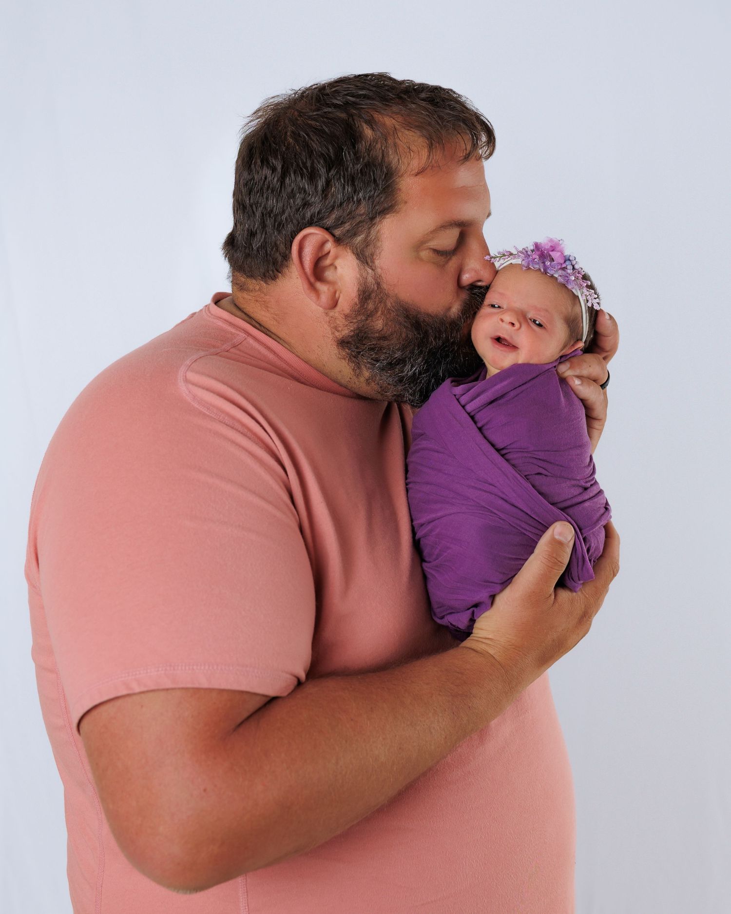 A tender moment shared between a bearded adult in a pink t-shirt and a newborn wrapped in purple swaddle with floral headband.