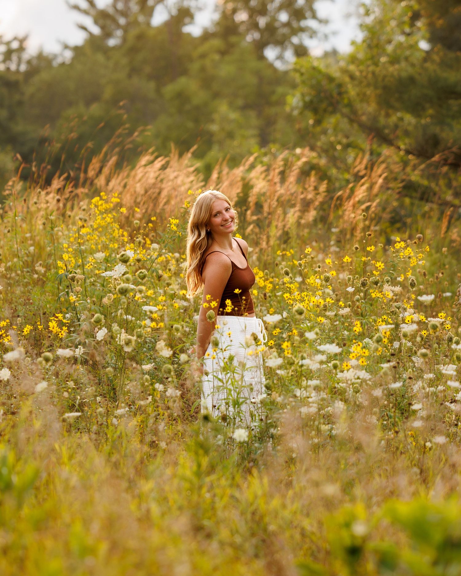 Young woman in white skirt and brown top smiling in a wildflower meadow with yellow and white blooms.