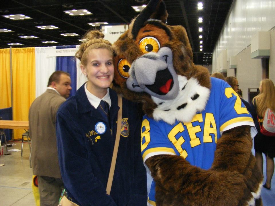 A school mascot in a brown bear costume poses with students in navy and gold uniforms in a hallway.