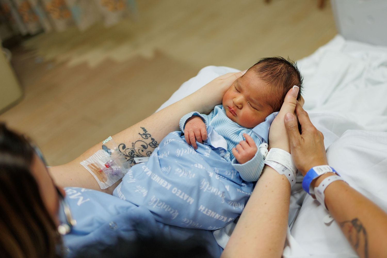 Newborn baby in blue outfit resting peacefully in hospital bed after birth, held by tattooed mother.