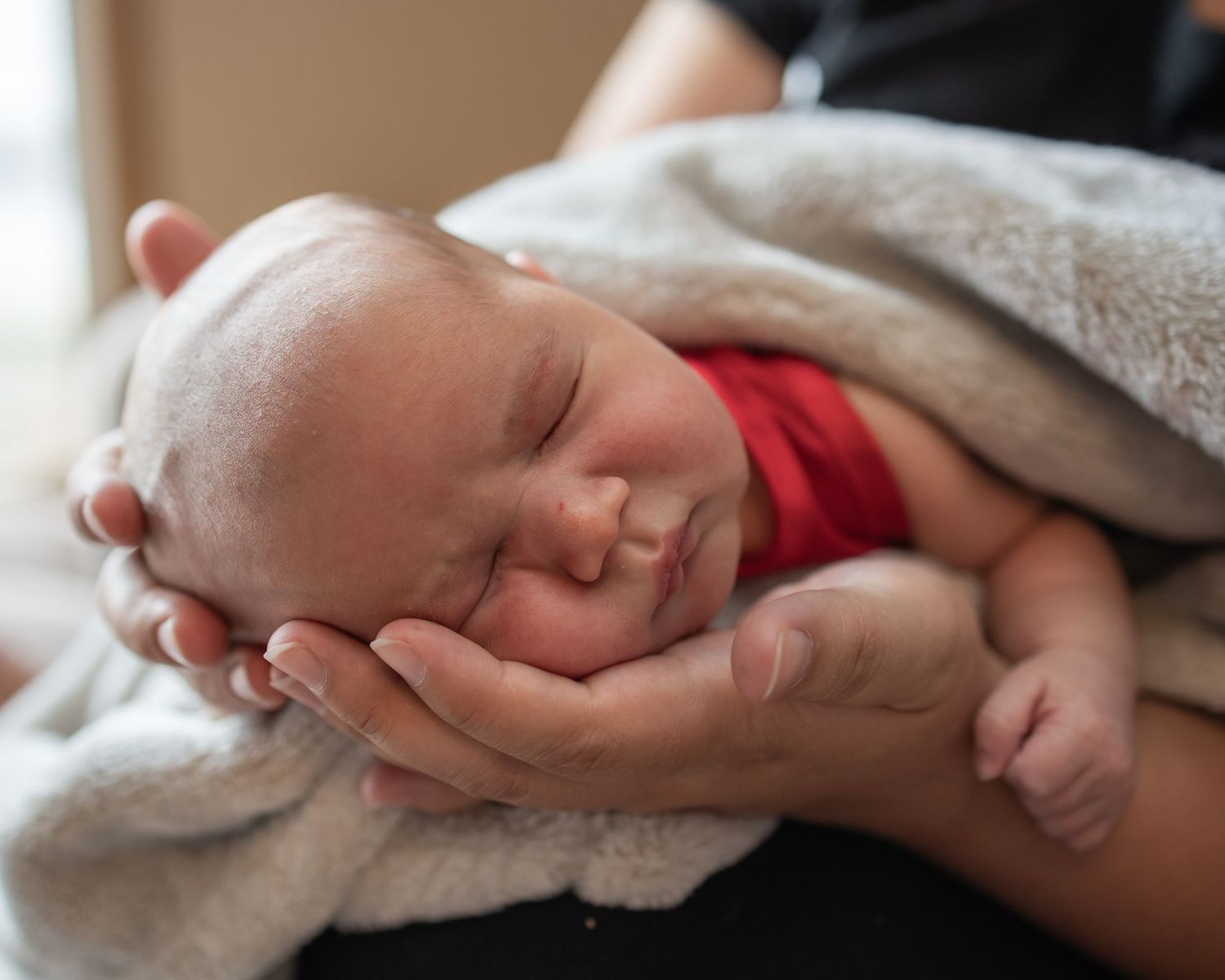 Peaceful sleeping newborn wearing red clothing cradled gently in hands under a soft gray blanket.