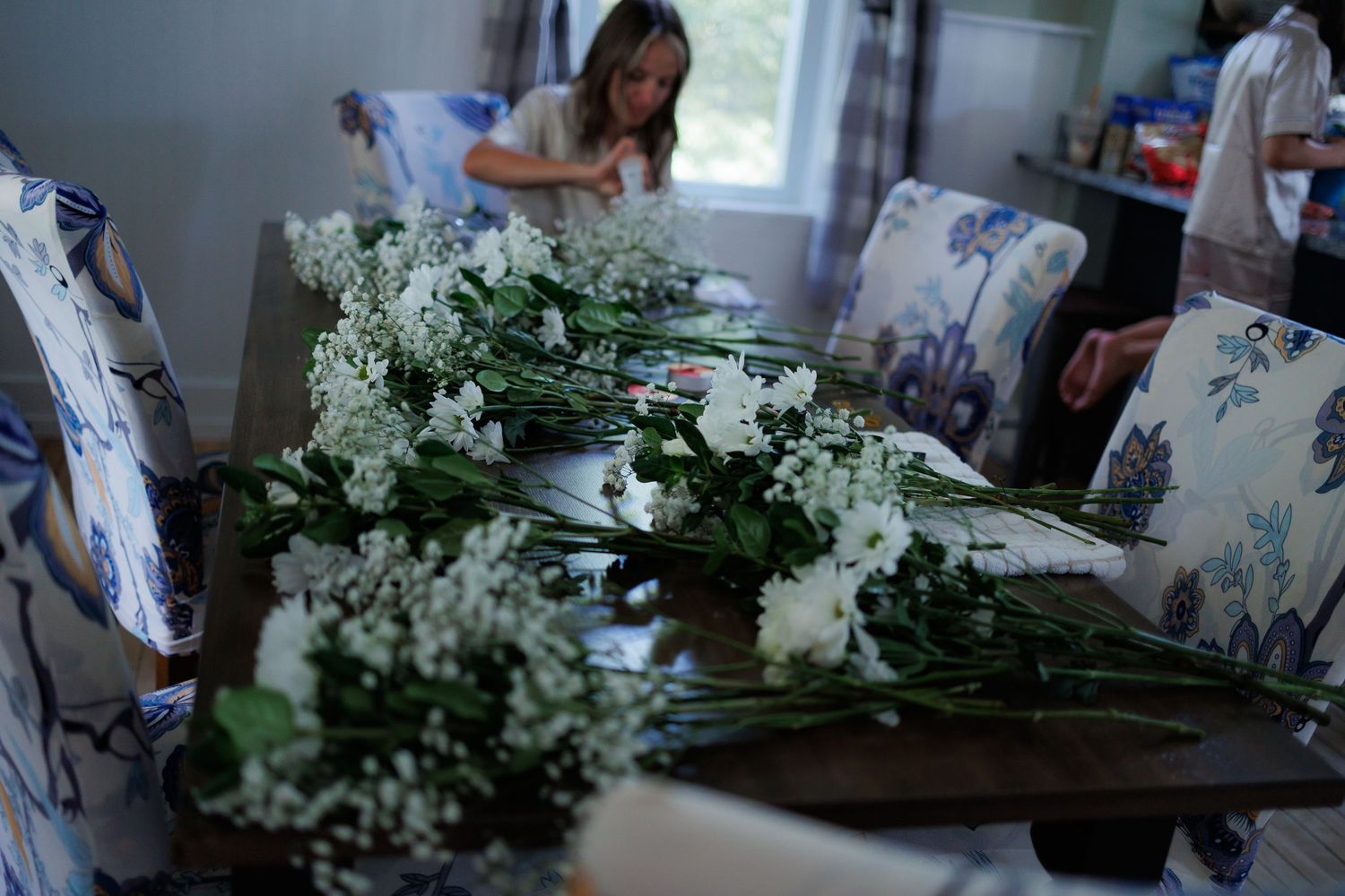 A person works with fresh white flowers and greenery at a floral design station in a bright workshop space.