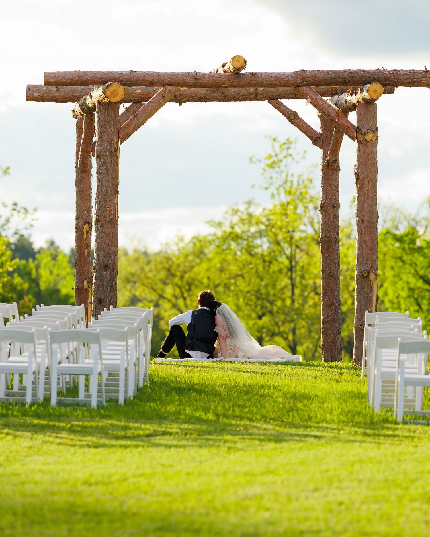 Rustic wooden wedding arch with mason jar lights overlooks rows of white chairs on a lush green lawn at sunset.
