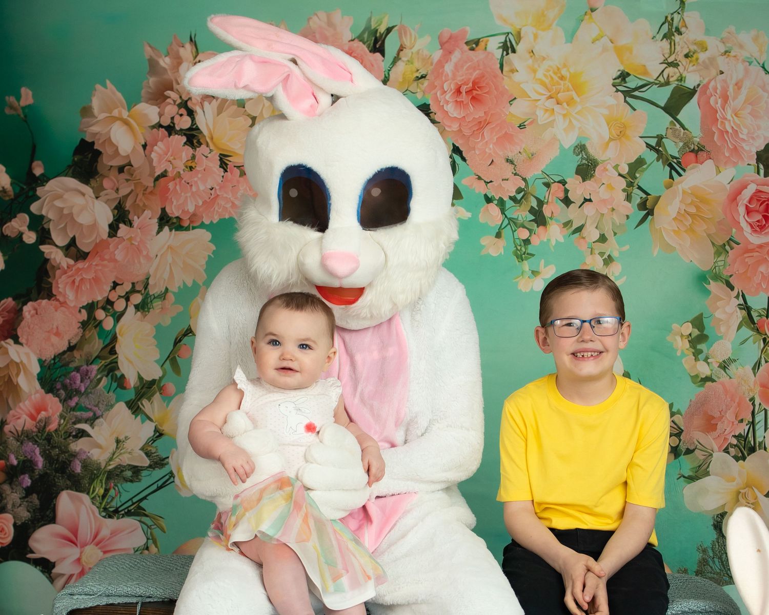 Portrait with Easter bunny costume figure seated between child in yellow shirt and baby in white dress against floral backdrop.