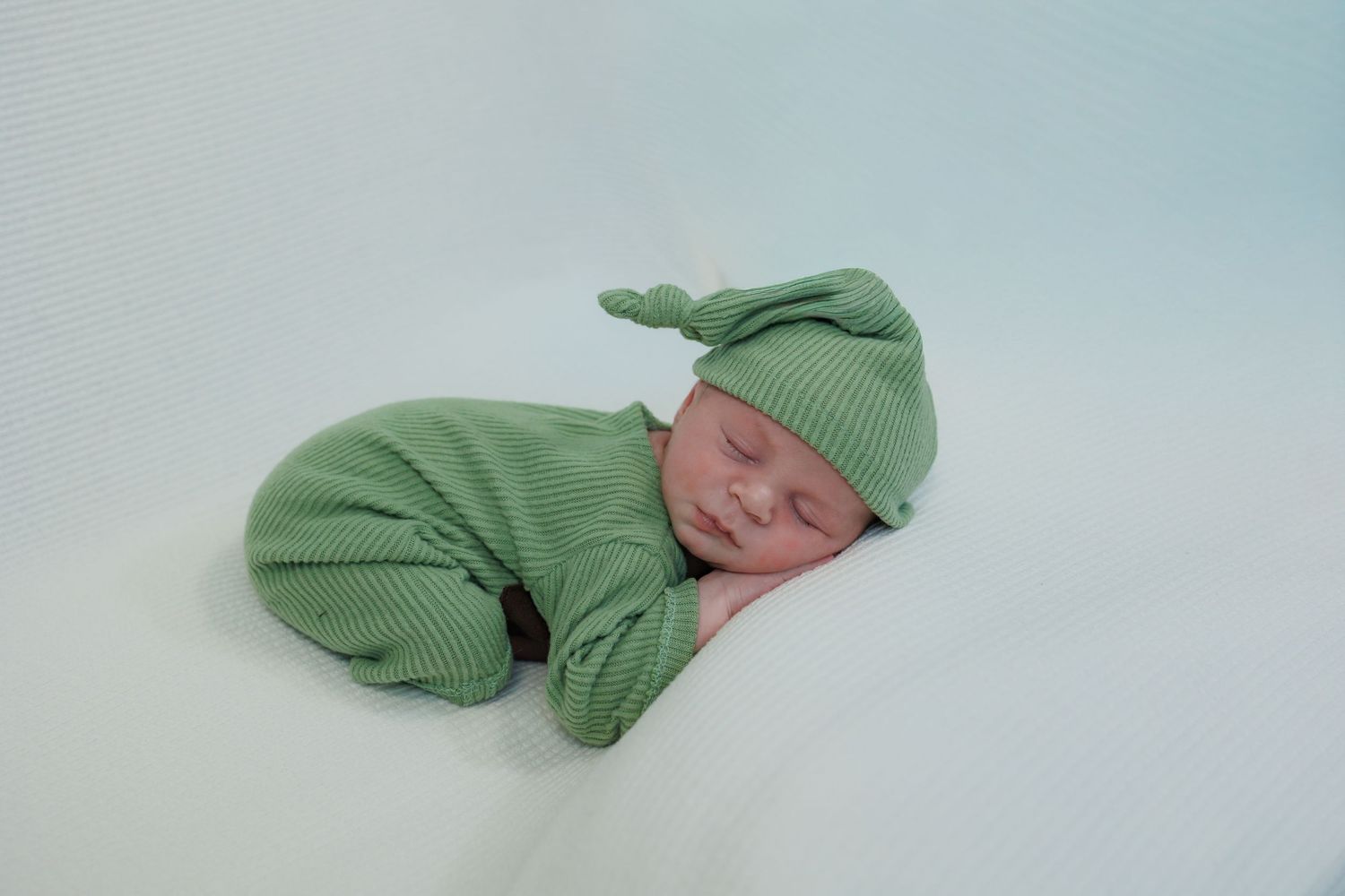 A sleeping newborn baby wearing green clothing and a knit hat rests peacefully on a soft grey blanket.