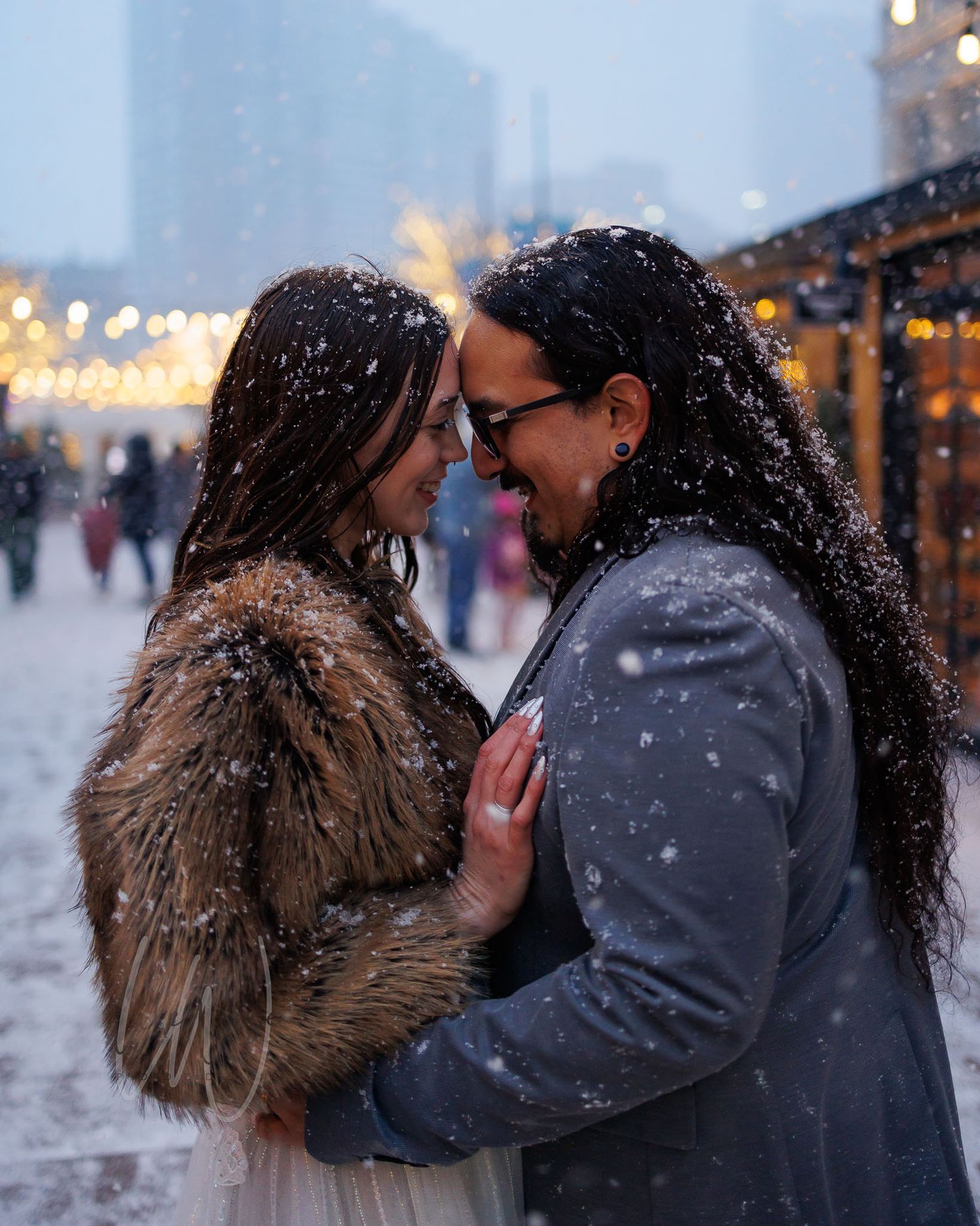 A romantic couple shares an intimate moment in the falling snow with twinkling city lights in the background.