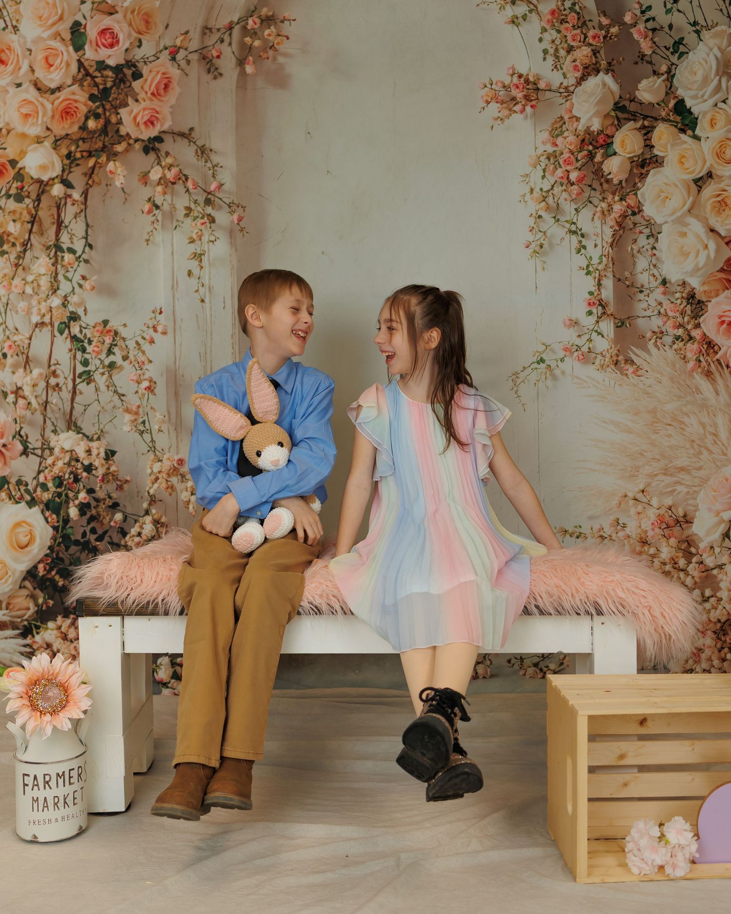 Two children sit together on a bench with floral decorations and pink feathers, sharing a sweet moment in a studio setting.